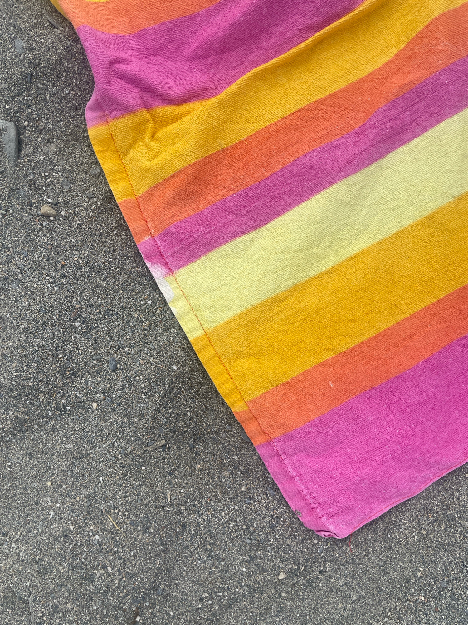 A striped towel, featuring pink, orange, yellow, and light yellow bands, lies on a dark, pebbly surface. The towel's edge is folded over on the bottom right. The scene appears to be a beach or promenade in La Herradura, Spain.