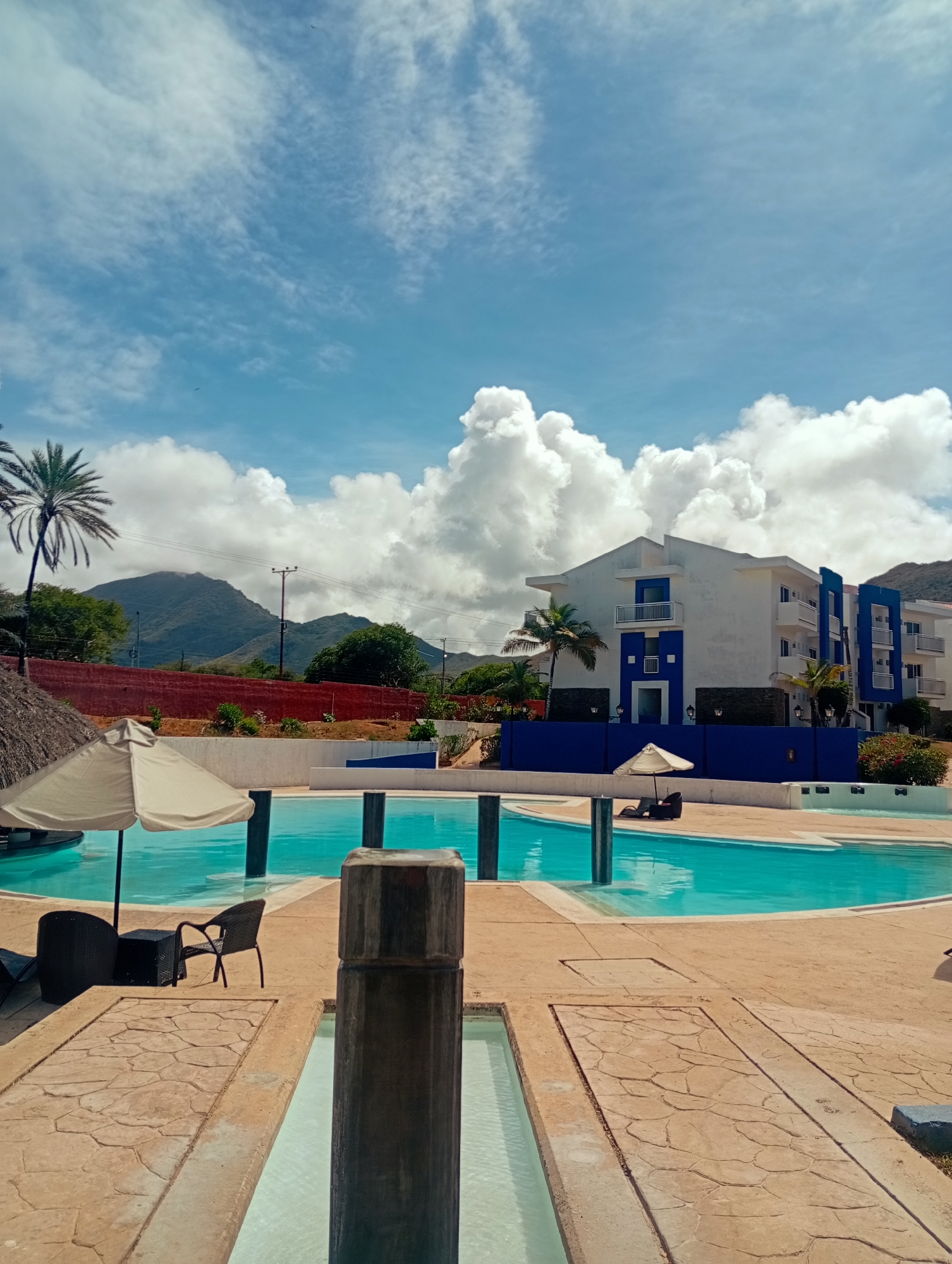 The image depicts a serene scene in Playa El Agua, Venezuela, featuring a multi-tiered swimming pool in the foreground, with water reflecting the bright, partly cloudy sky above. The pool is surrounded by a light-brown, patterned concrete area. Lounge chairs and umbrellas are strategically placed around the pool. In the background, a modern, white and blue building stands behind a dark blue wall, with palm trees scattered around it. Further in the background, rolling hills and mountains are visible. The overall impression is one of a peaceful resort setting on a sunny day.