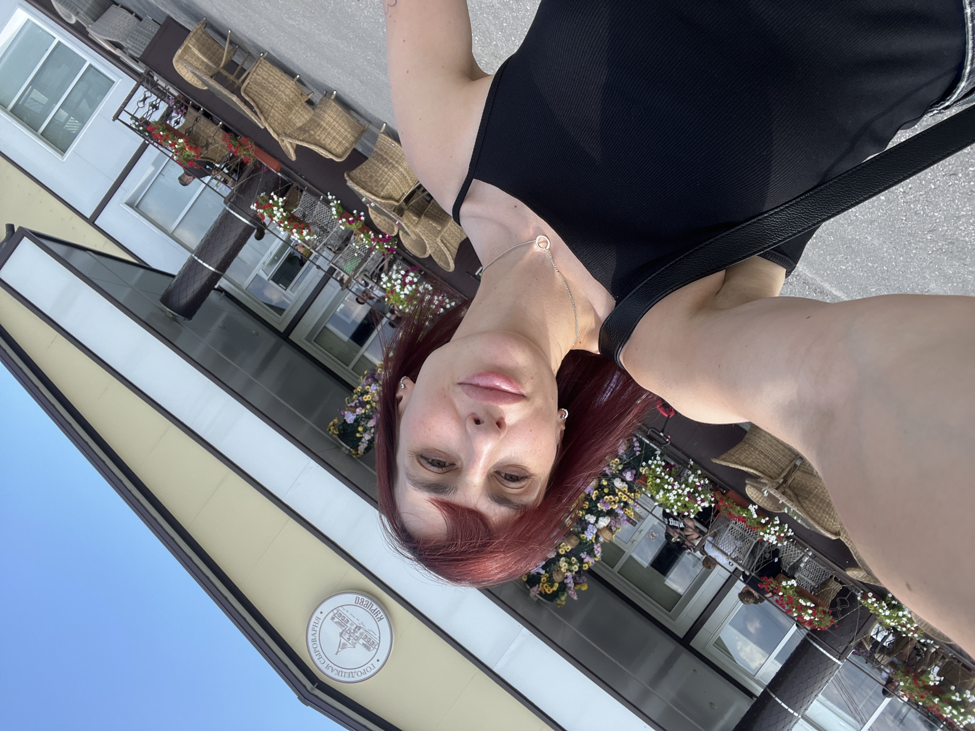 A woman with red hair takes a selfie in front of a building that appears to be a restaurant or cafe. The building has a light-colored facade with brown trim and windows. There are outdoor seating areas with wicker chairs and tables, some of which are occupied by people. Flower boxes filled with red and white flowers adorn the balcony and facade. A circular sign with text and an image of a building is mounted on the upper part of the facade. The sky is clear and blue. The location is identified as Akhlebaikha (Kovriginskii S/S), Russia.