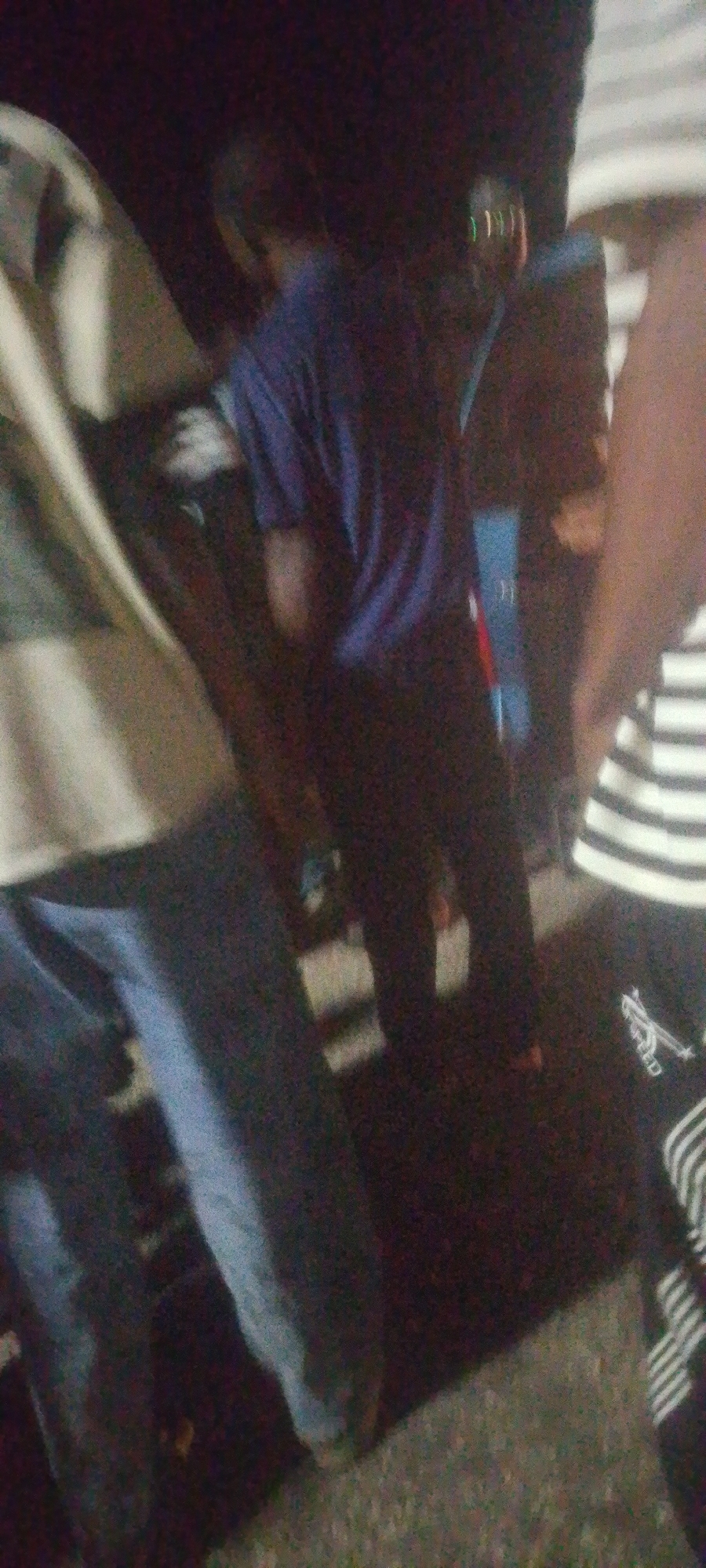 A group of people stands on a paved surface in Adankolo, Nigeria. One person wears a blue t-shirt and dark pants and appears to be barefoot. Another individual is wearing a striped shirt. Dim lighting conditions prevail. A partial view of jeans is also visible on the left side of the frame.