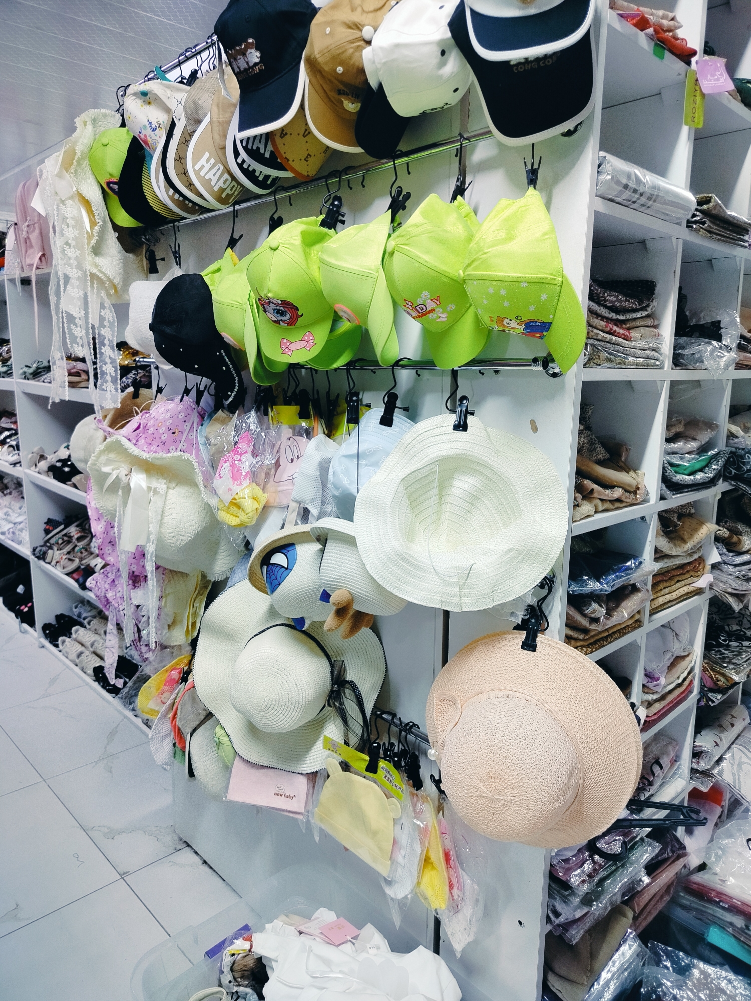 This is a medium shot photograph of a retail store display, likely a children's clothing or accessories shop, in the city of Kölkent, Kazakhstan. The primary focus is a wall-mounted display of various hats and caps hanging from metal racks. The hats are diverse in style and color, including baseball caps, sun hats, and novelty hats, some with cartoon characters or bright patterns.
To the right of the hat display, shelves are filled with folded clothing items, predominantly in pastel colors and intricate patterns. Some items are individually packaged in plastic. The flooring appears to be tiled in a light, marbled pattern. The overall impression is one of an organized retail environment, with a focus on children's apparel. There are no people visible in the image.