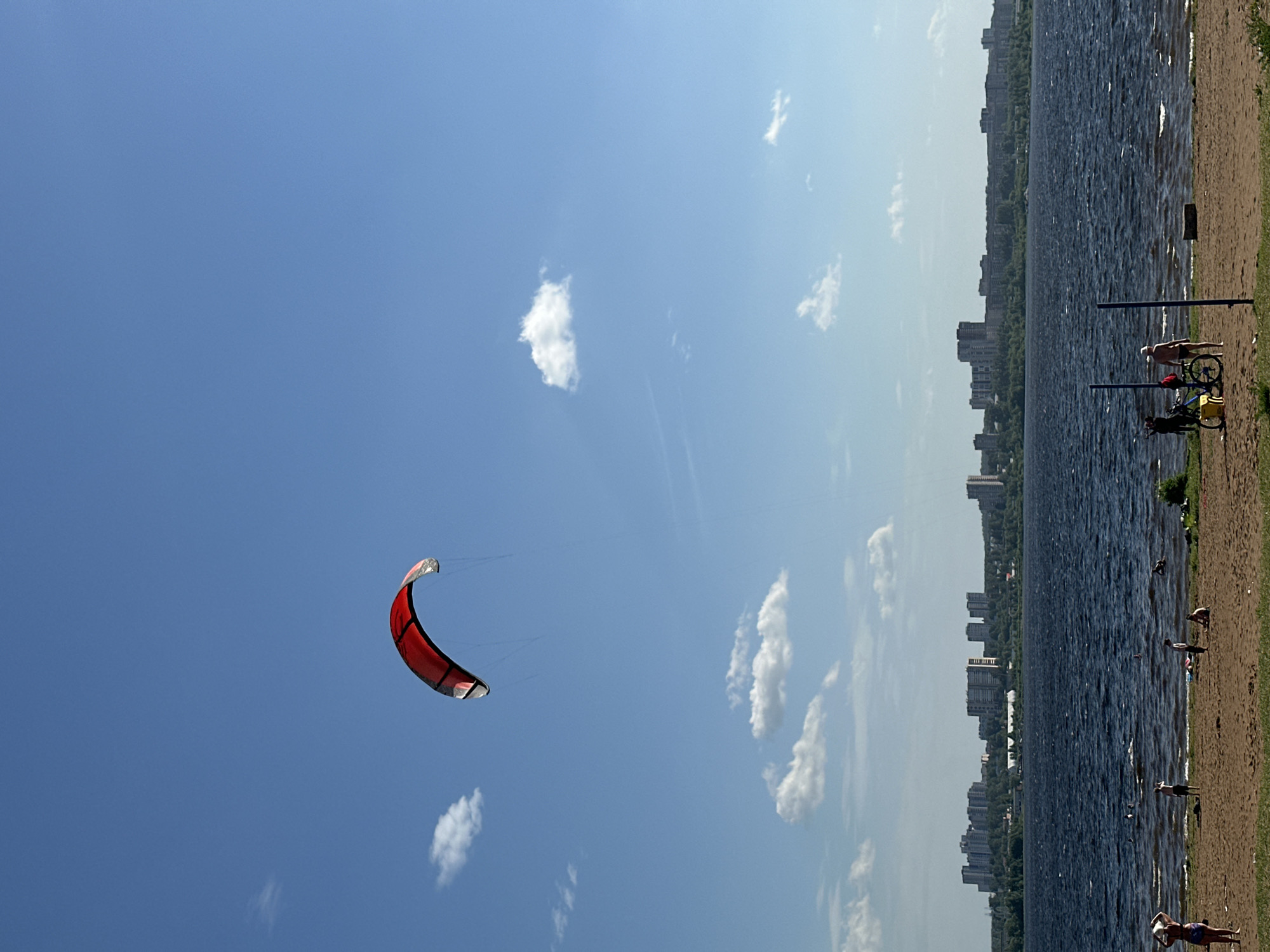 The image shows a kitesurfing kite in the sky over a body of water, likely a lake or reservoir, near Izhevsk, Russia. The kite is red and black, positioned in the upper left portion of the frame. The sky is a clear blue with scattered cumulus clouds. The body of water takes up the right half of the image, with a shoreline visible on the right edge of the frame. The shoreline is a sandy beach with a grassy area behind it. People are present on the beach, some standing and some lying down. A bicycle is parked near the water's edge. In the background, across the body of water, are buildings that are characteristic of a city skyline. Based on the cityscape and the presence of the large body of water, this is likely the Izhevsk reservoir in Izhevsk, Russia.