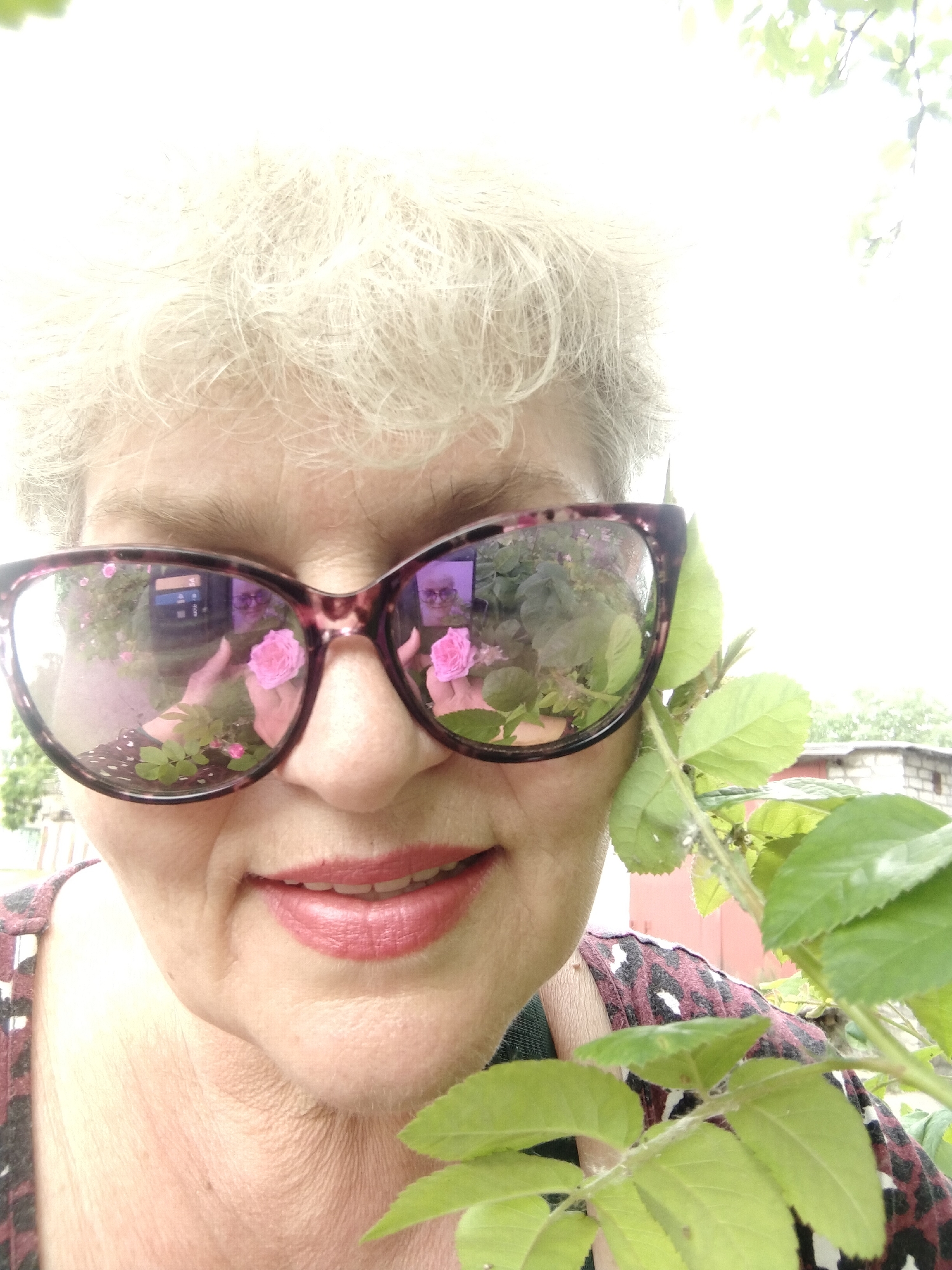 A bright, close-up outdoor portrait, likely a selfie, features an older woman with short, curly gray hair, smiling subtly at the viewer. She is wearing large sunglasses with mottled brown-purple frames. The lenses provide a clear reflection of the scene in front of her, showing her hands holding a vibrant pink rose, with smaller green leaves visible around it. A faint reflection of the woman's face and possibly the camera she's using is also visible within the sunglasses. She is dressed in a dark top with an abstract or animal-print pattern in shades of maroon, black, and white. To the right side of her face, a branch with fresh green rose leaves is prominently positioned, partially framing her and indicating a natural, garden-like setting. The lighting is strong and bright, suggesting a sunny daytime. In the background, beyond the foliage, a light-colored building, possibly a brick outbuilding or shed, with a darker roof is visible. The bright sky and distant green trees complete the outdoor scene. No discernible text is visible in the image. The setting is likely Horishni Plavni, Ukraine.