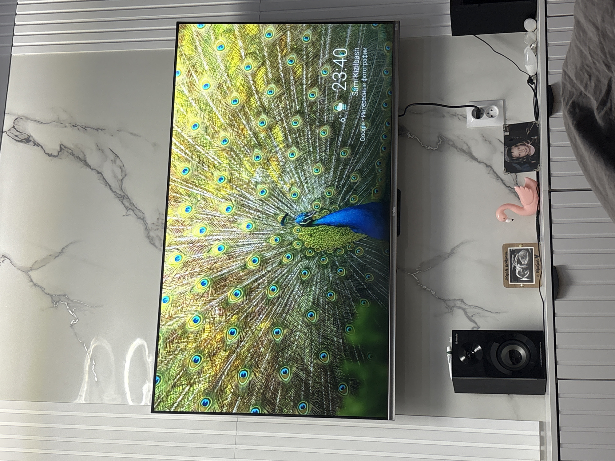 The image shows a television mounted on a wall with a peacock feather pattern displayed on the screen. The time on the screen indicates 23:40, with weather information showing "6°," and the name "Sami Kizilbash" along with "Google • Интересные фотографии" (Google • Interesting photos) written in Russian. There is a power outlet, a photograph, a small pink flamingo figurine, and another small framed image affixed to the wall adjacent to the television. A black speaker is mounted below these items. The wall itself has a marble-like pattern and is bordered by horizontally placed white tiles above and below. The location is indicated as Chaikovsky, Russia, based on the information displayed on the television screen. A portion of a person wearing gray clothing is visible in the upper right corner.