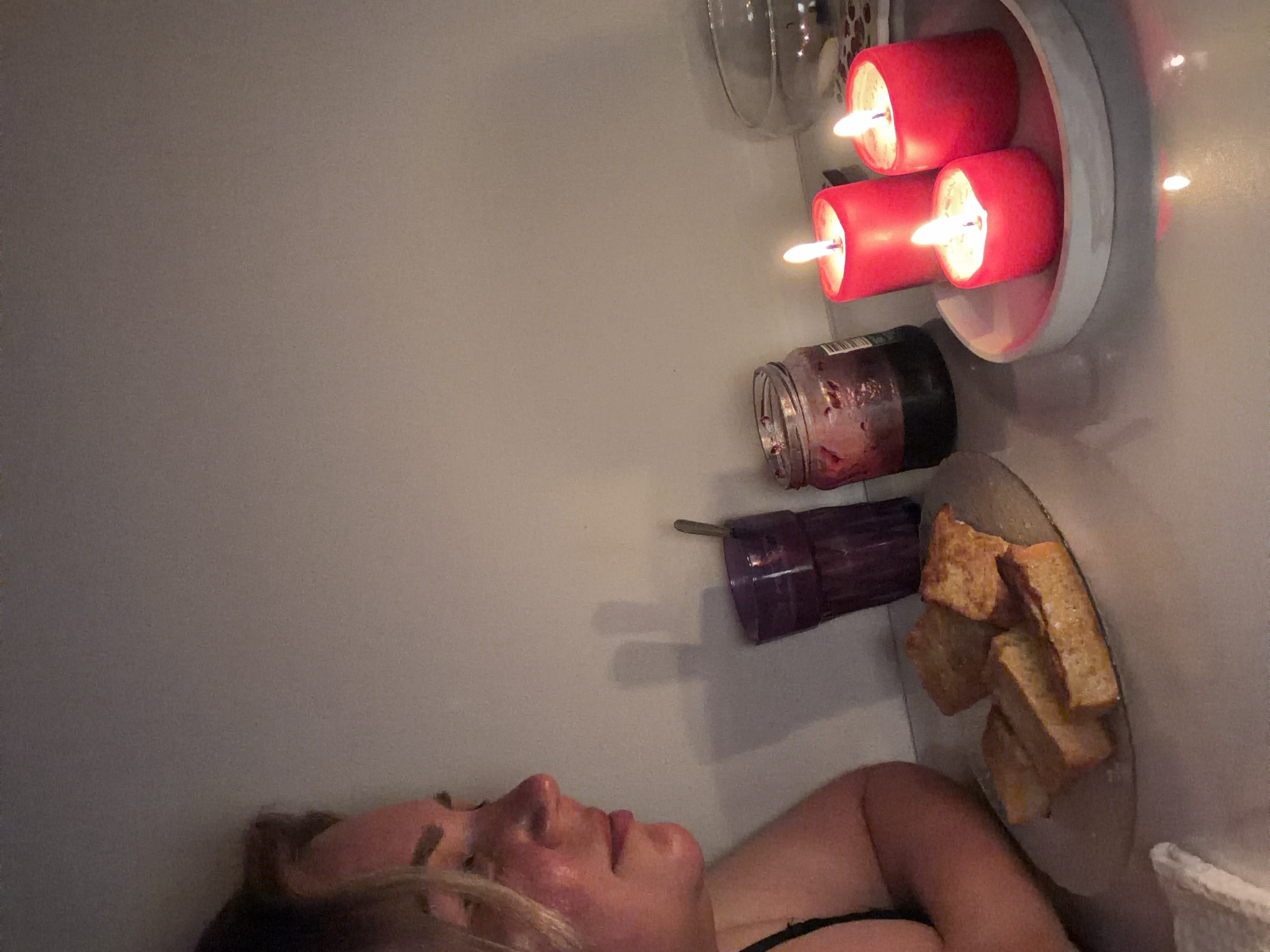 The image depicts a person lying in a bathtub. Next to them are several objects: a plate with pieces of toast, a purple glass, a jar, and a dish with three lit red candles. The background is a plain wall. The location, Leningradskaya, Russia, is not determinable from the image alone.