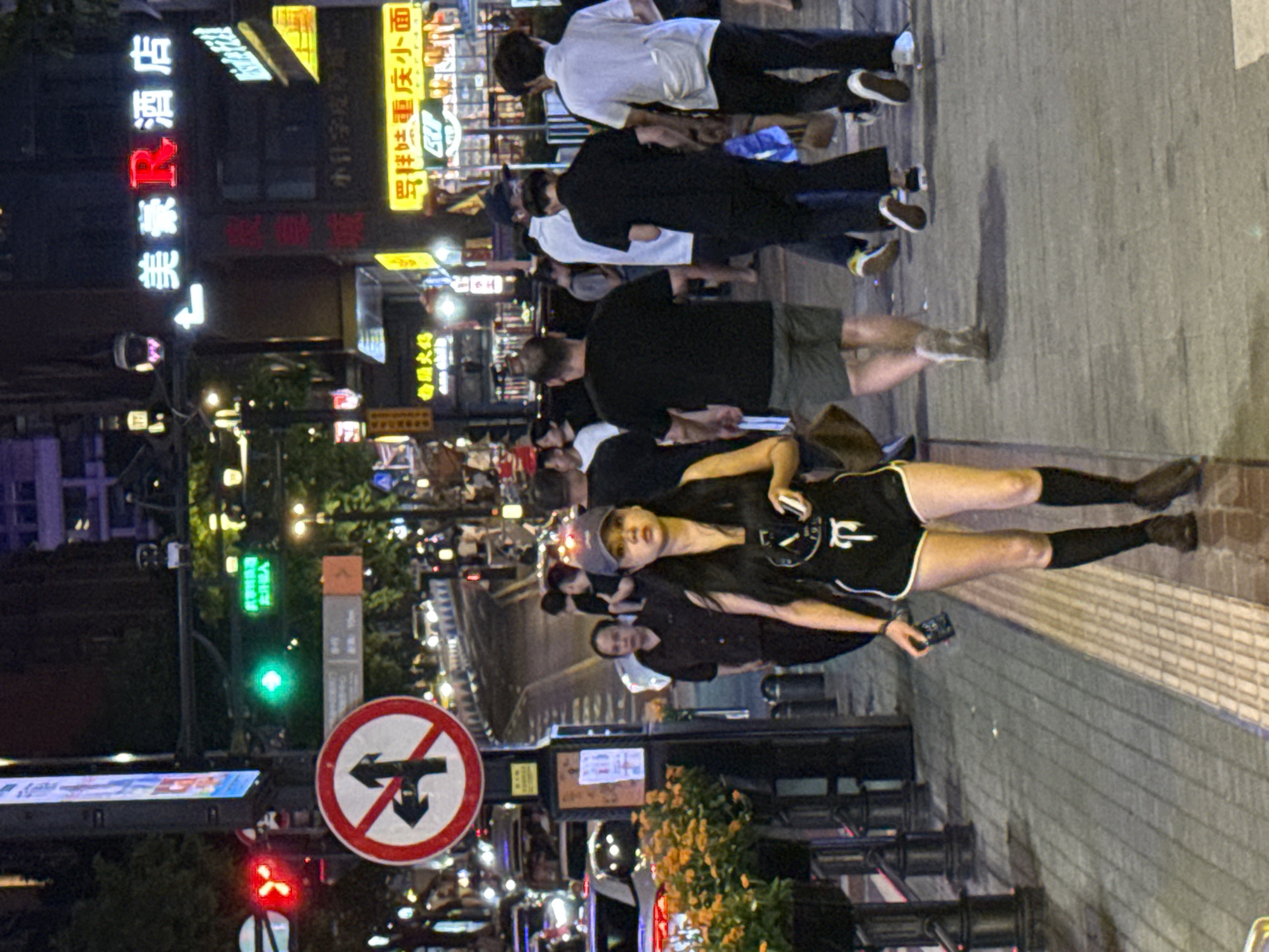 The image shows a street scene at night in Yuzhong District, China. A young woman in athletic attire, black shorts with white trim, a black sleeveless top, and knee-high black socks, walks towards the camera, holding a smartphone in her right hand. She is wearing a grey baseball cap. Behind her, several people are walking in various directions. To the left of the frame, a traffic light displays a red signal, and a circular sign with a red border and a diagonal line indicates "no left or straight turn." A green traffic light is visible further up the street, as well as neon signs for businesses, including "美衆R酒店" (Meizhong R Hotel). The street is paved with interlocking stones, and there are planters with orange flowers on the left. The environment is illuminated by streetlights and the glowing signs of commercial establishments.