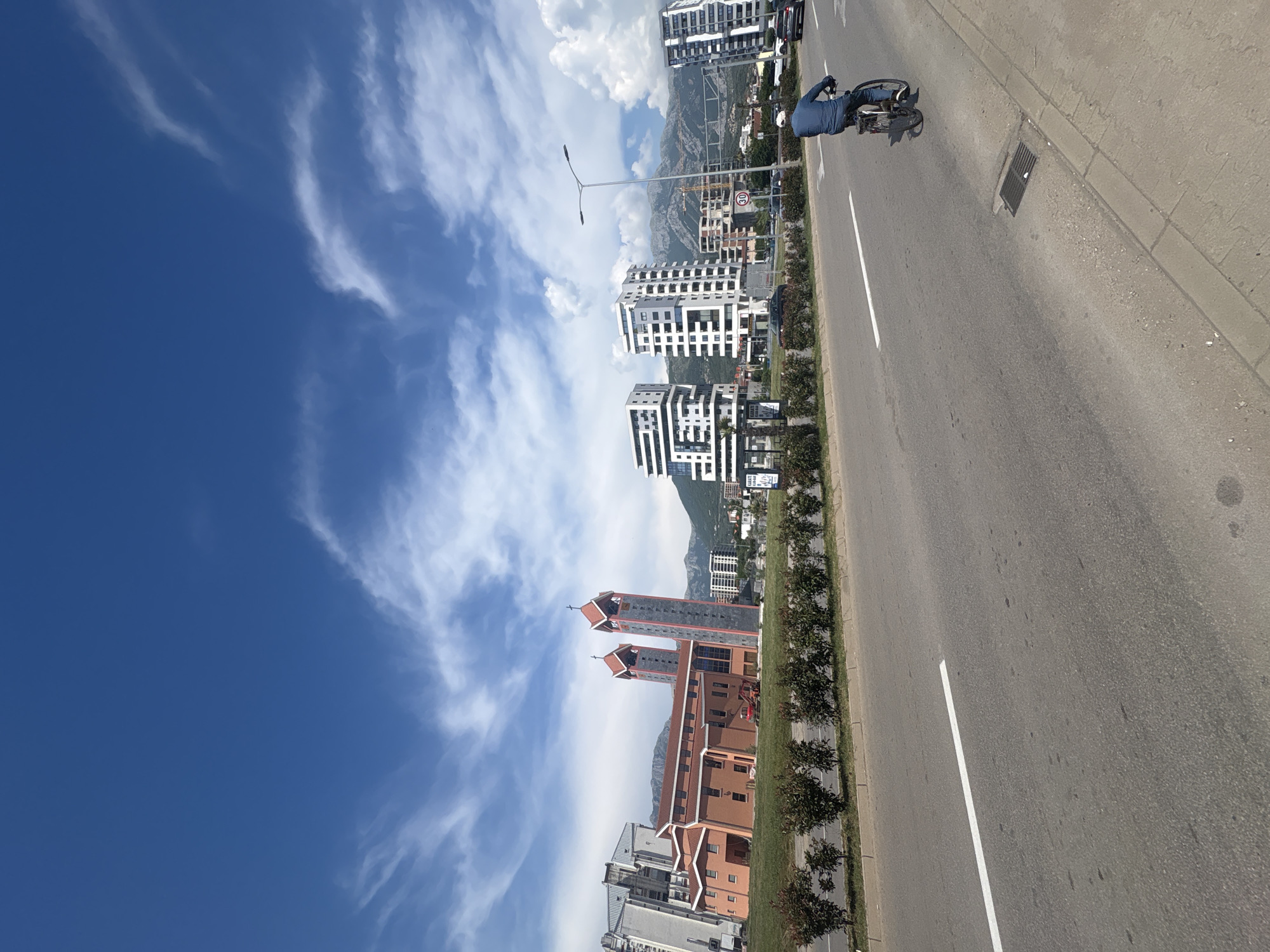 This image captures a street scene in Bar, Montenegro, on a bright, sunny day with a partly cloudy blue sky. In the foreground, a man on a bicycle rides along the right side of a paved road marked with white lines. The road has a curb and sidewalk to its right. Behind the road, a row of manicured bushes lines a grassy area.
Further back, a striking red brick church with two tall, gray bell towers topped with red roofs and crosses is prominently visible. To the left of the church, a tall, modern apartment building stands. In the middle ground and to the right of the church, several contemporary white high-rise apartment buildings rise against a backdrop of rolling, verdant mountains. Construction cranes are also visible among the buildings. The overall impression is of a developing urban area in a scenic coastal or mountainous region.