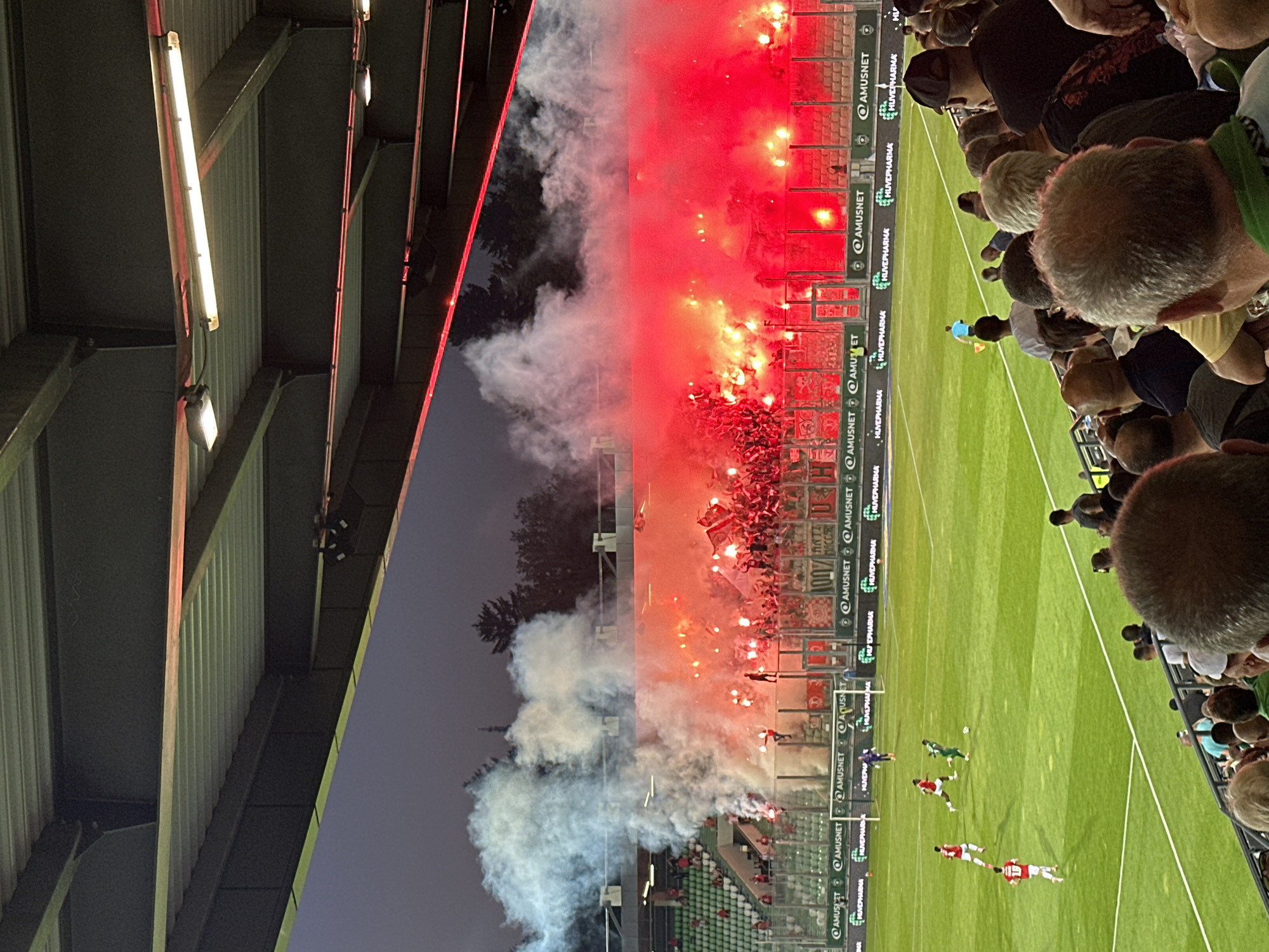 The image depicts a football match at the Huvepharma Arena in Razgrad, Bulgaria. The scene is characterized by intense fan activity, with a large number of supporters in the stands releasing red and white flares and smoke, creating a dramatic atmosphere. The pitch is illuminated by floodlights, and a football game is in progress with players in red and green kits. Spectators are visible in the foreground and background, observing the match and the pyrotechnics. The stadium infrastructure, including seating areas and a canopy, is also visible. The sky is dark, indicating it is likely evening or night.