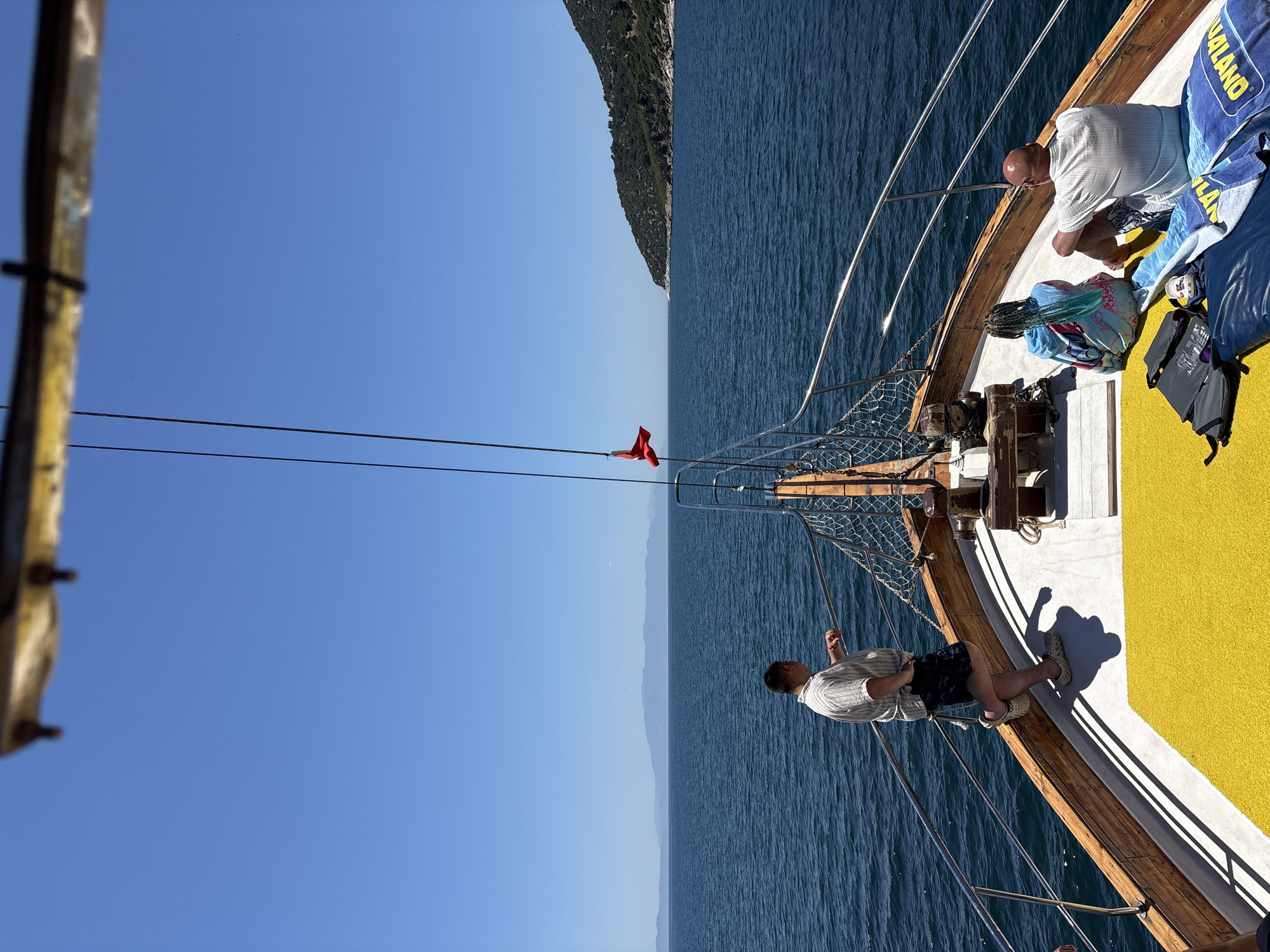 The image captures a vibrant daytime scene aboard a boat, likely a tour boat, sailing on calm blue waters under a clear, bright blue sky. A prominent, green mountainous coastline stretches along the distant horizon, indicating a coastal setting.
On the boat's deck, which features warm wooden planks and a bright yellow non-slip surface, several individuals are present. Towards the right side of the frame, a bald man in a white striped shirt is seen crouching, looking down towards the water. Nearby, a person with distinctive braided hair is seated, wrapped in a patterned blanket or towel. Blue towels are also visible on the deck, one partially displaying the text "AQUALAND." Further towards the bow, on the left, another man in a light-colored striped shirt and dark shorts stands, leaning against the railing and looking out to sea.
A small red flag flutters from the boat's rigging at the front. The strong sunlight indicates it is midday or early afternoon, casting distinct shadows from the people and railings onto the yellow deck. The scene conveys a relaxed, leisurely atmosphere typical of a holiday excursion. Given the mountainous coast and calm waters, the setting is consistent with the provided location context of Menderes, Turkey.