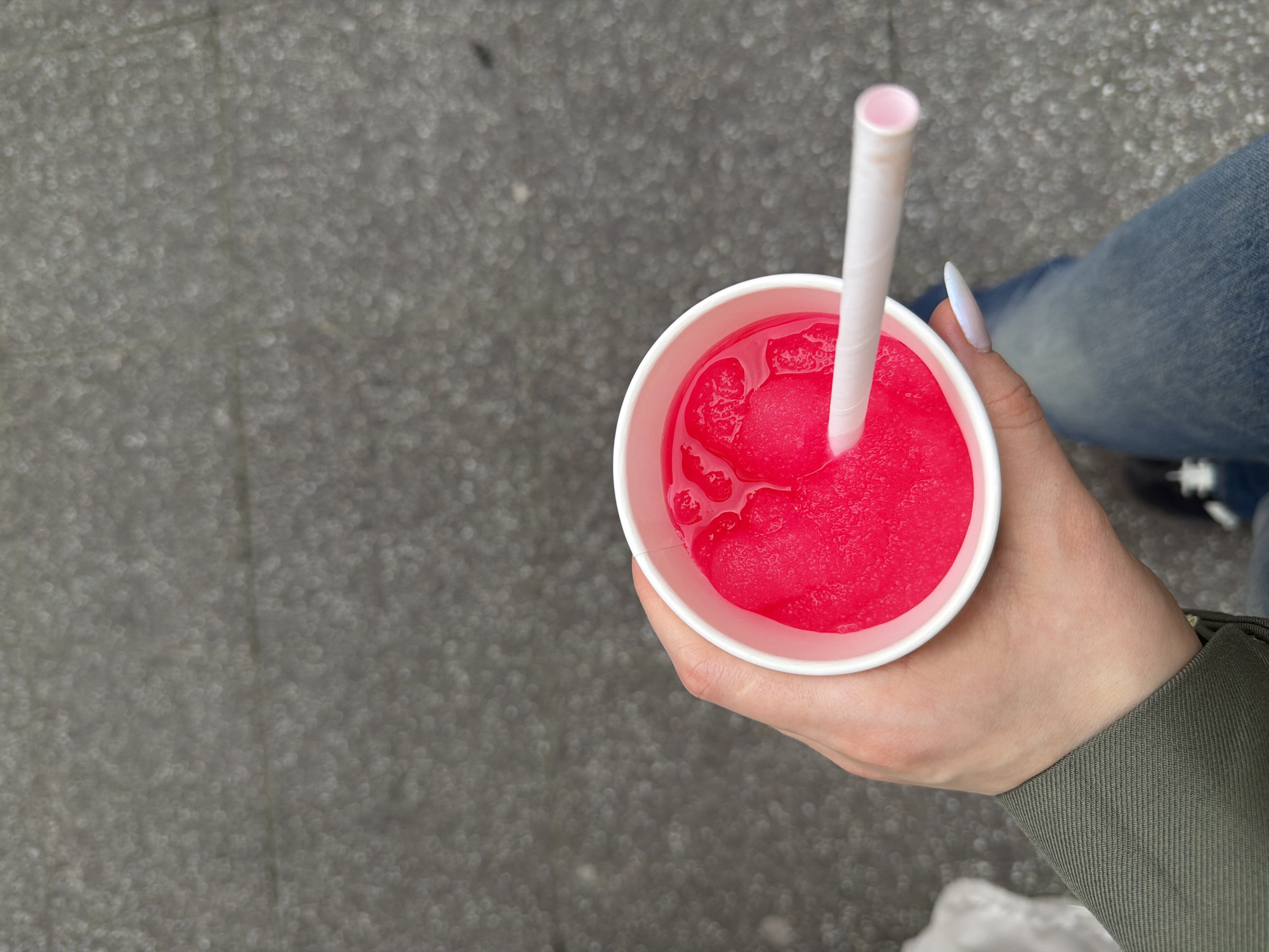 A high-angle medium shot shows a person holding a cup of red slushy with a white straw in their right hand. The person is standing outside on a gray stone-textured surface, possibly a sidewalk. The cup is white and contains a bright red slushy drink. A white straw is inserted in the drink. The person has a fair complexion and white nail polish. They are wearing a light green jacket and jeans. Based on the architectural style of the background and overall ambiance, the location is presumed to be the city of Strasbourg, France.