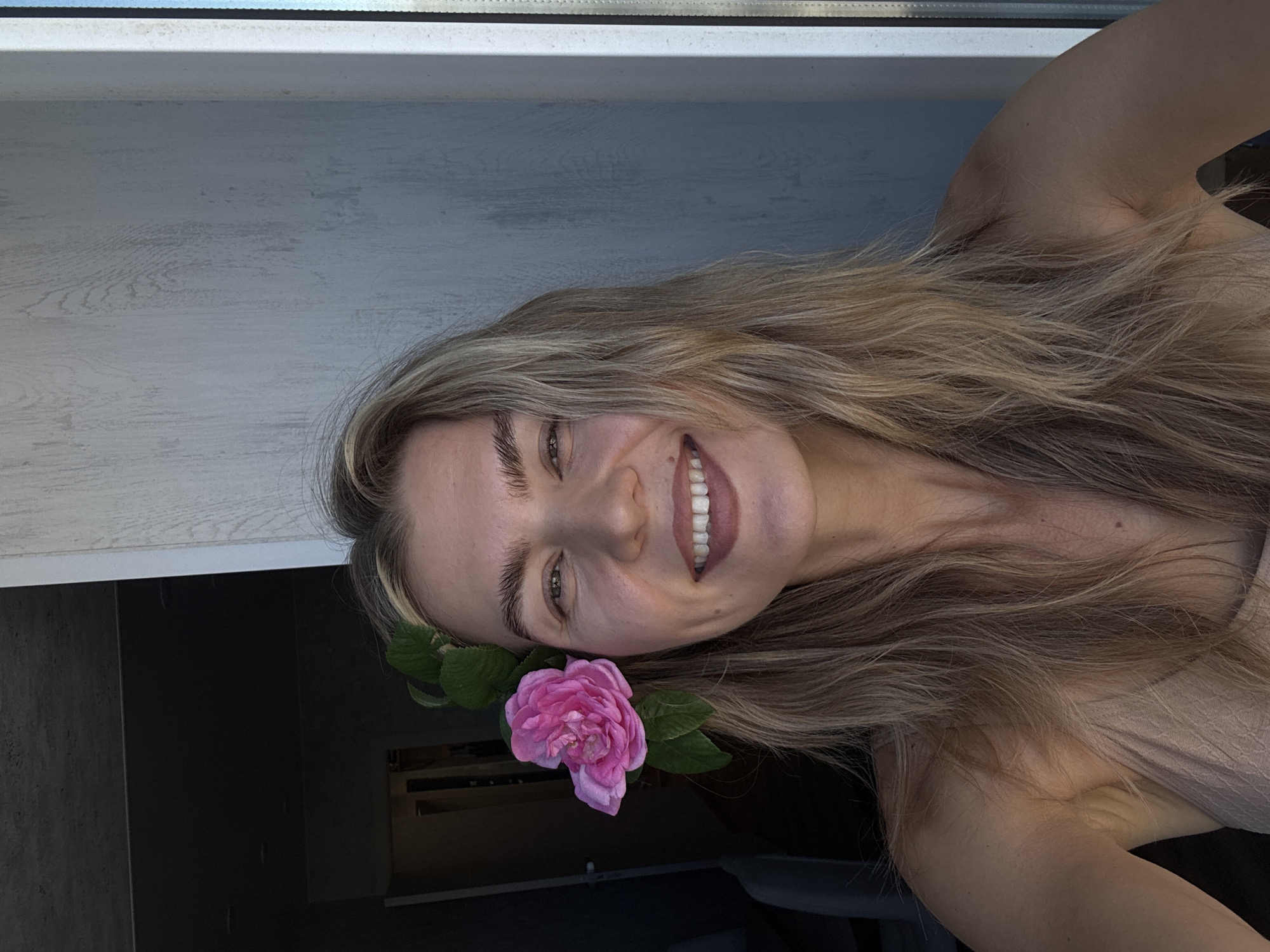 The image is presented rotated 90 degrees clockwise. It features a young woman smiling broadly, her face and upper body positioned centrally within the frame. She has long, wavy blonde and light brown hair that flows towards the right side of the rotated image. A vibrant pink rose with several green leaves is tucked into her hair, visible near what would be her right ear if the image were upright. Her expression is joyful and relaxed, with a wide smile revealing white teeth. Her light-colored skin is visible on her face, shoulders, and arms, and she appears to be wearing a light-colored top.
In the background, positioned above her head in the rotated image, a white window frame is visible. Below this, a light-colored, textured wall with a subtle wood grain pattern occupies a significant portion of the background. The lower left portion of the background is darker, appearing as a black or dark gray surface. The scene is brightly lit, suggesting it is daytime indoors. No text is visible in the image. The overall mood conveyed is cheerful and friendly.