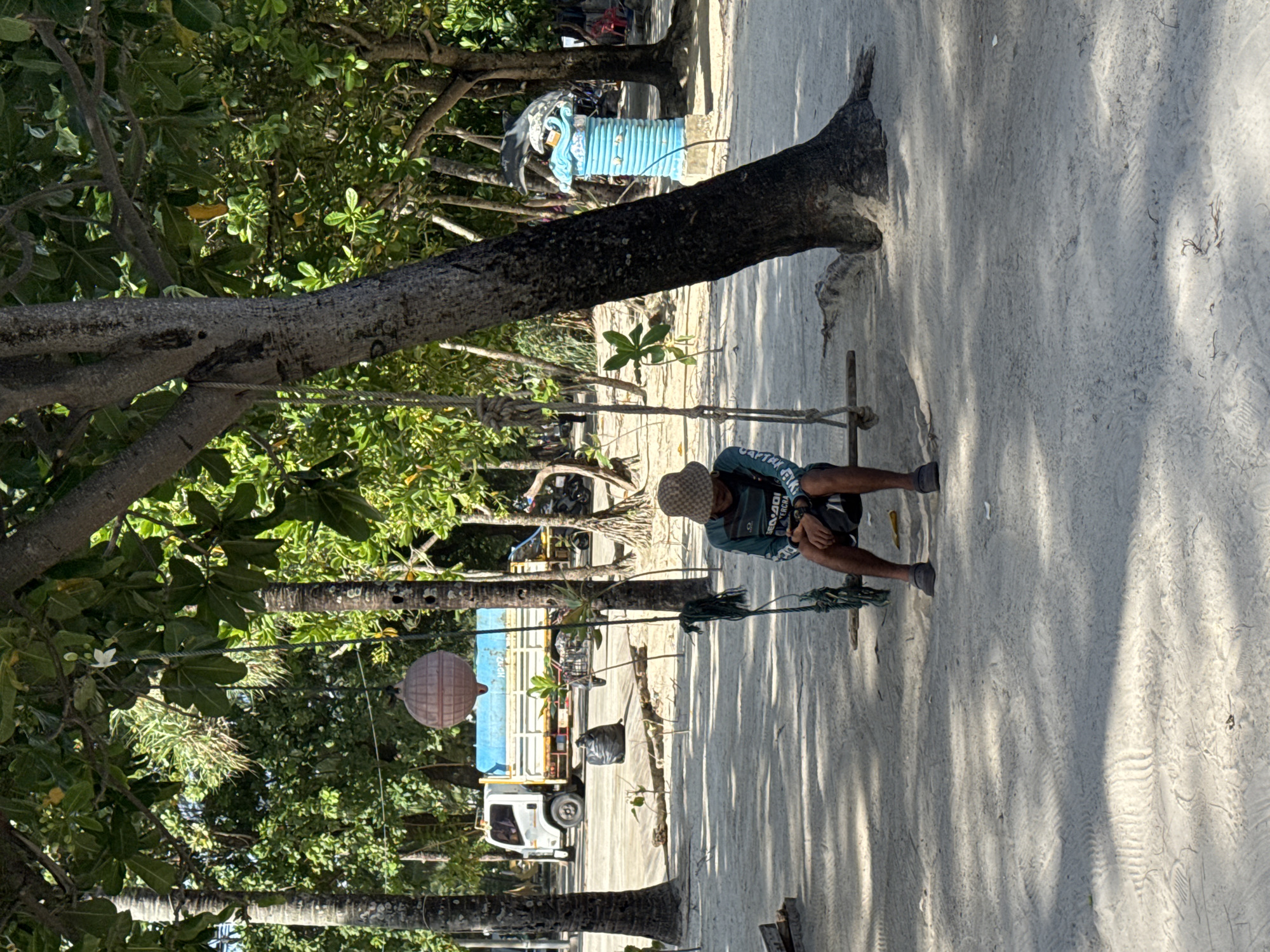 In Patong, Thailand, a person sits on a wooden swing suspended by ropes from a tree. The individual, wearing a patterned bucket hat, a long-sleeved teal shirt with white lettering, dark shorts, and dark shoes, is looking down at a dark object in their hands. The swing hangs in a sandy area under the shade of trees with dense green foliage. In the background, a white truck with a blue tarp-covered bed is parked. To the right of the truck is a black garbage bag. Further in the background, a decorative blue pillar with a dolphin on top is visible. Several other trees and some distant structures can be seen, creating a tropical outdoor environment.