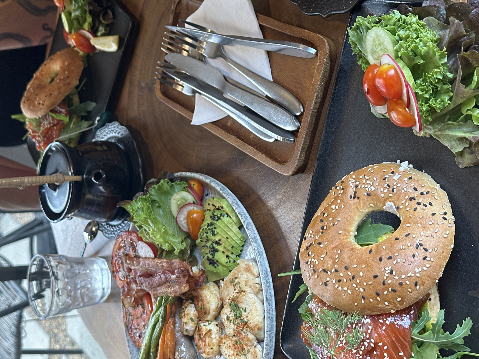 A still life food scene at Baan Bo Phut, Thailand. A sesame bagel sandwich is on a black plate with greens. Another bagel sandwich is on a dark plate. A silver tray with avocado, meat, green beans, and other vegetables is visible. A wooden tray holds silverware. A metal coffee or tea dispenser is visible. A glass is partially visible on the table. The environment suggests a casual restaurant or cafe setting.