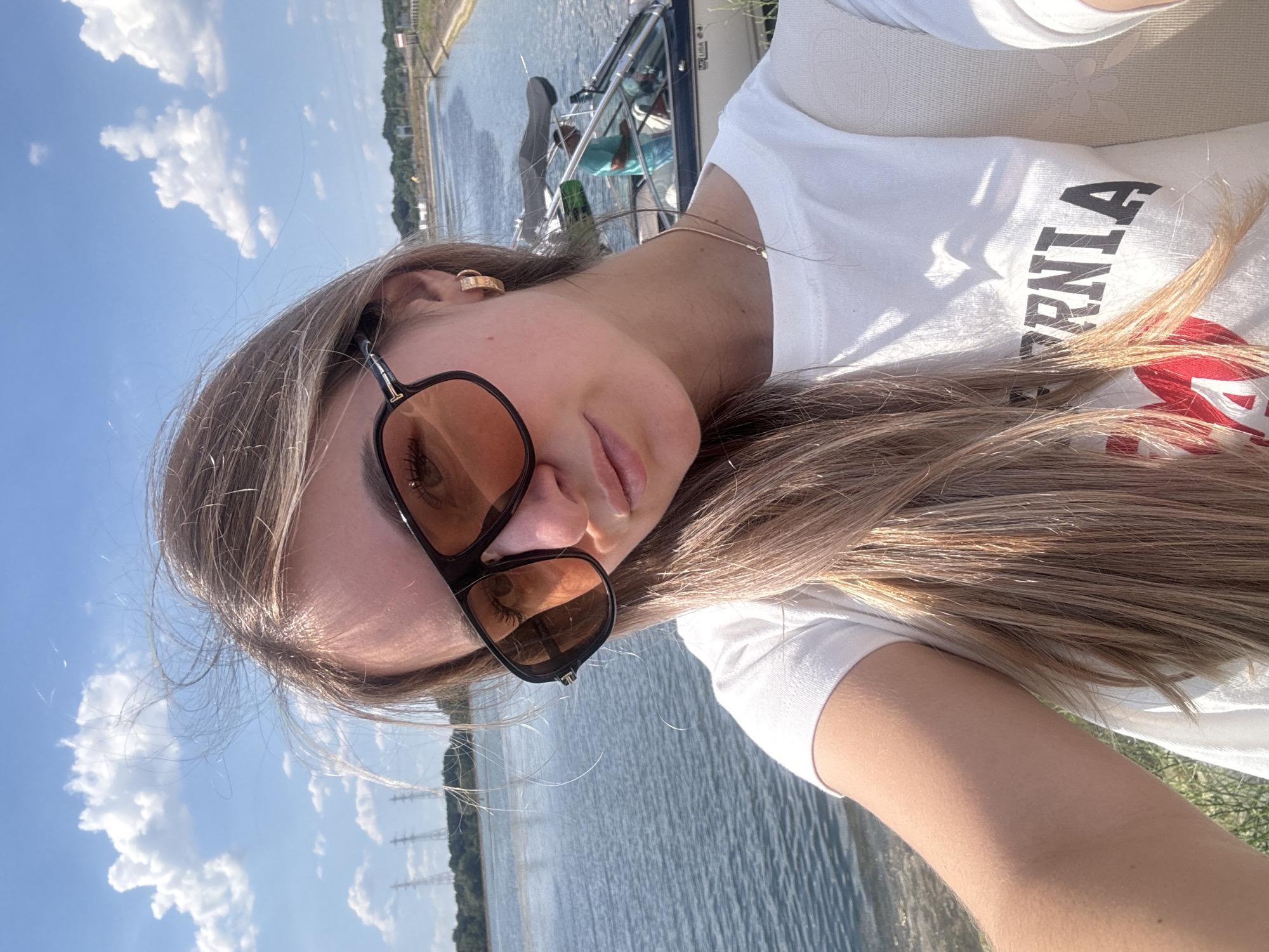 A young woman with blonde hair, wearing sunglasses and a white t-shirt, takes a selfie outdoors. She is positioned in the foreground, with a body of water, a boat with a person in it, and trees in the background. The sky is blue with white clouds. The woman is wearing a gold hoop earring. The t-shirt has "CALIFORNIA" and a red heart graphic visible. The scene appears to be a waterfront area.