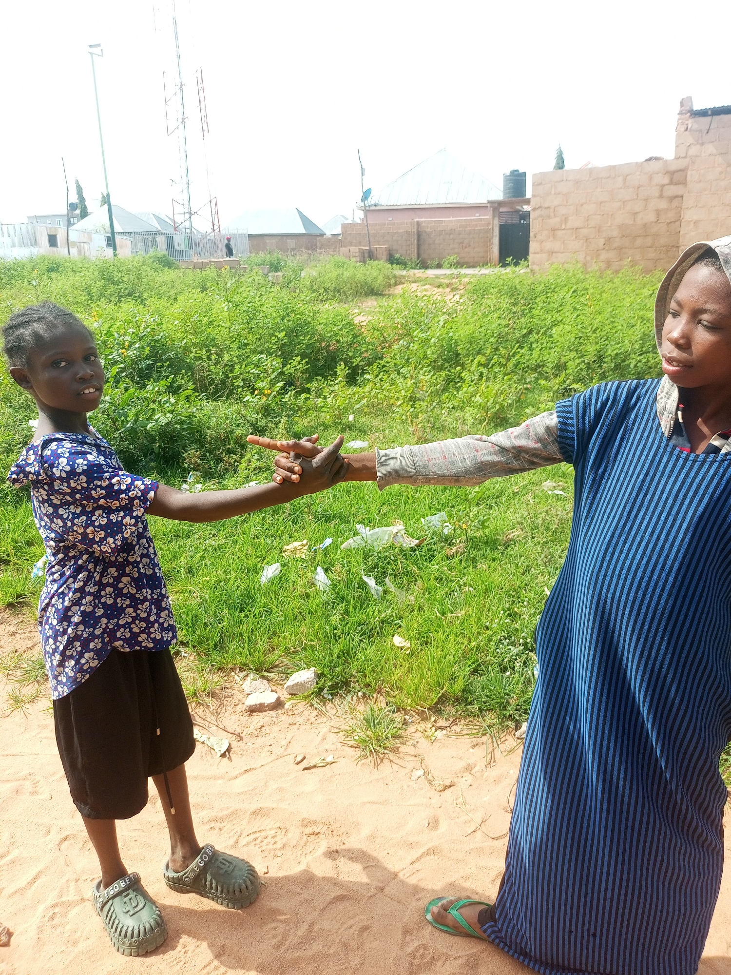 Two young girls are standing on a sandy path in Tempure, Nigeria, with their hands clasped. The girl on the left, wearing a blue and white floral print dress and black shorts, looks at the camera with her left hand extended, fingers pointing. The girl on the right, dressed in a blue striped dress with a hood, is looking away from the camera and is holding the other girl's hand. They are surrounded by green grass and scattered litter. In the background, there are buildings, a telecommunications tower, and a brick wall. The sky is bright and overcast.