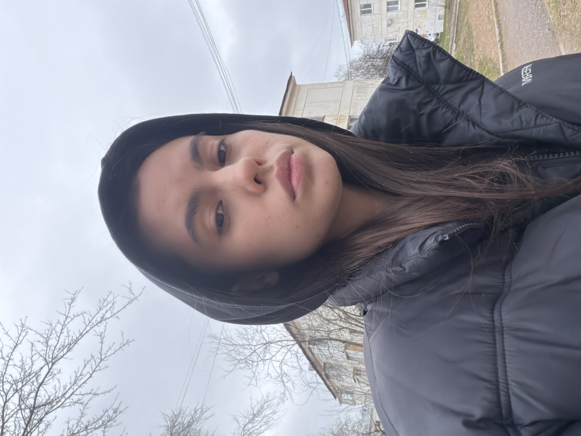 A low-angle, close-up self-portrait captures a young woman outdoors in Chaikovsky, Russia. The image is taken from below, looking up towards the overcast sky. The woman occupies the majority of the frame, positioned slightly off-center. She has long, dark brown hair that falls over her shoulders, and fair skin. Her expression is neutral to slightly serious as she looks directly into the camera. She is wearing a black, hooded puffer jacket with a zipper partially visible. A distinctive detail is partially visible white text on the right shoulder of her jacket (viewer's left, due to the photo's orientation), which appears to be mirrored and reads "ЯЕВЛЯЧ" (YEVLYACH) in Cyrillic script.
The background reveals a typical urban or suburban setting under a grey, cloudy sky, indicating overcast weather. Bare tree branches are prominent on the left and bottom right sides, suggesting late autumn, winter, or early spring. Several power lines or telephone wires stretch across the top of the frame. In the distance, parts of older residential buildings or apartment blocks, characterized by light-colored facades and visible windows, can be seen, reinforcing the Russian location context. The overall lighting is soft and diffuse due to the cloudy conditions, indicating daytime.