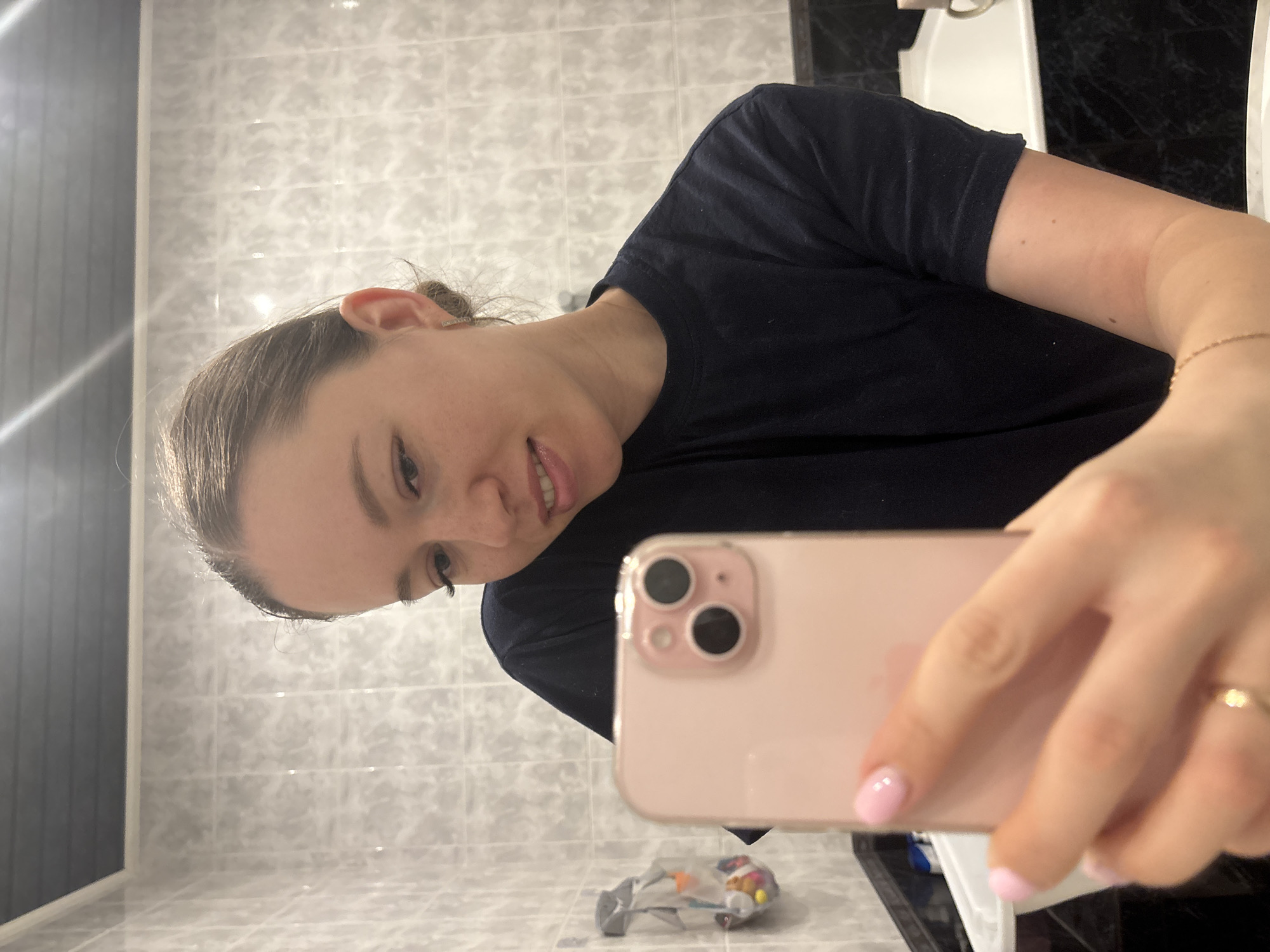 The image shows a person taking a selfie in a bathroom. The person, likely female, is wearing a dark blue t-shirt and has her hair pulled back. She is holding a pink iPhone. The bathroom has white and grey tiled walls. A white sink is visible in the background. There is also a small bag of items on the floor. I have no information about the location in the city of Ферма, Russia.
