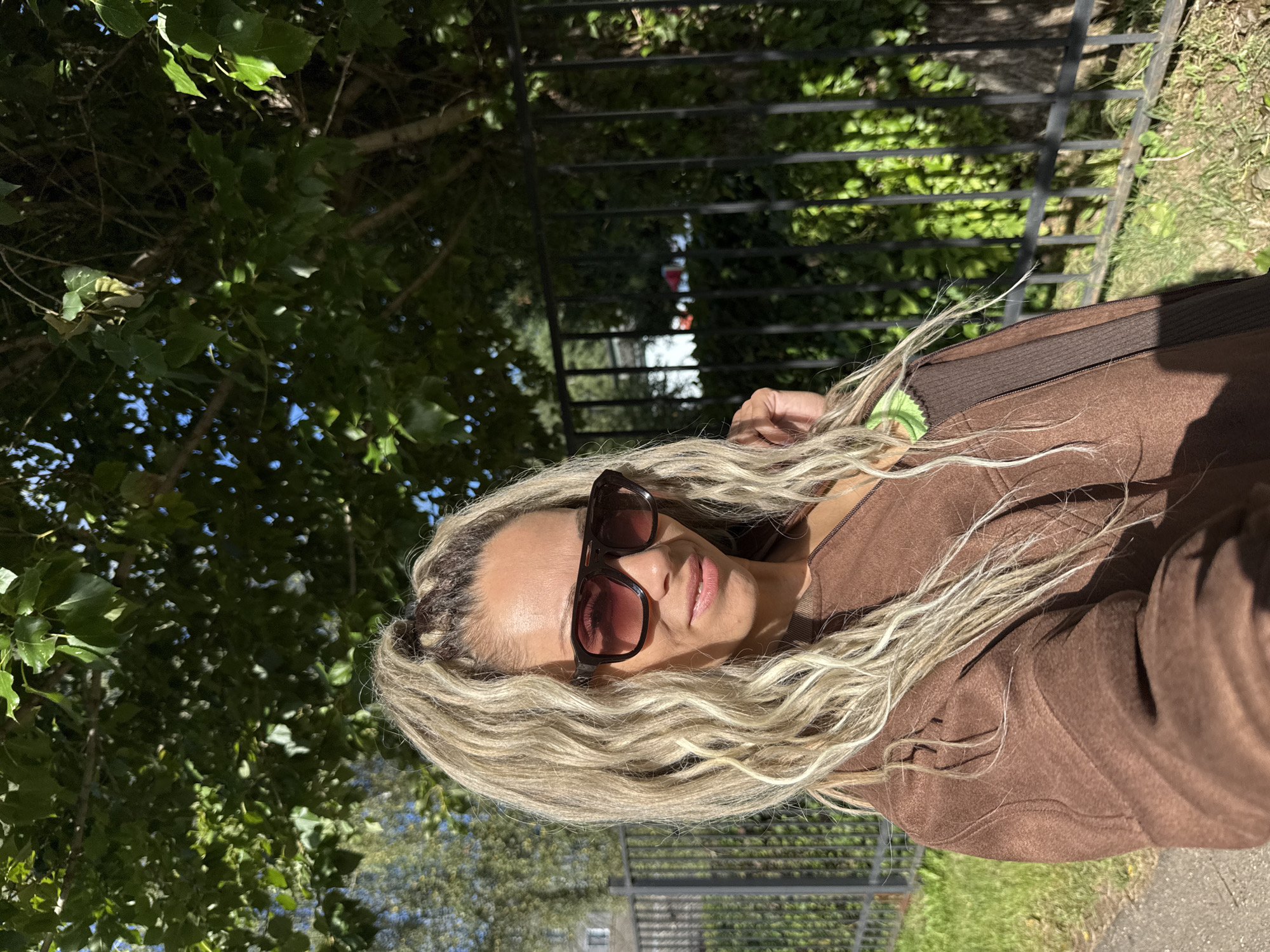 A woman with blonde dreadlocks is taking a selfie outdoors in front of a black metal fence. She is wearing large, dark-rimmed sunglasses and a brown zippered jacket. The background shows green trees and foliage, with a patch of blue sky visible through the leaves. The ground is paved, with grass and a sidewalk. The image appears to be taken on a sunny day. The provided location of Rusanovo, Russia, cannot be confirmed or denied based on the visual information in the image.