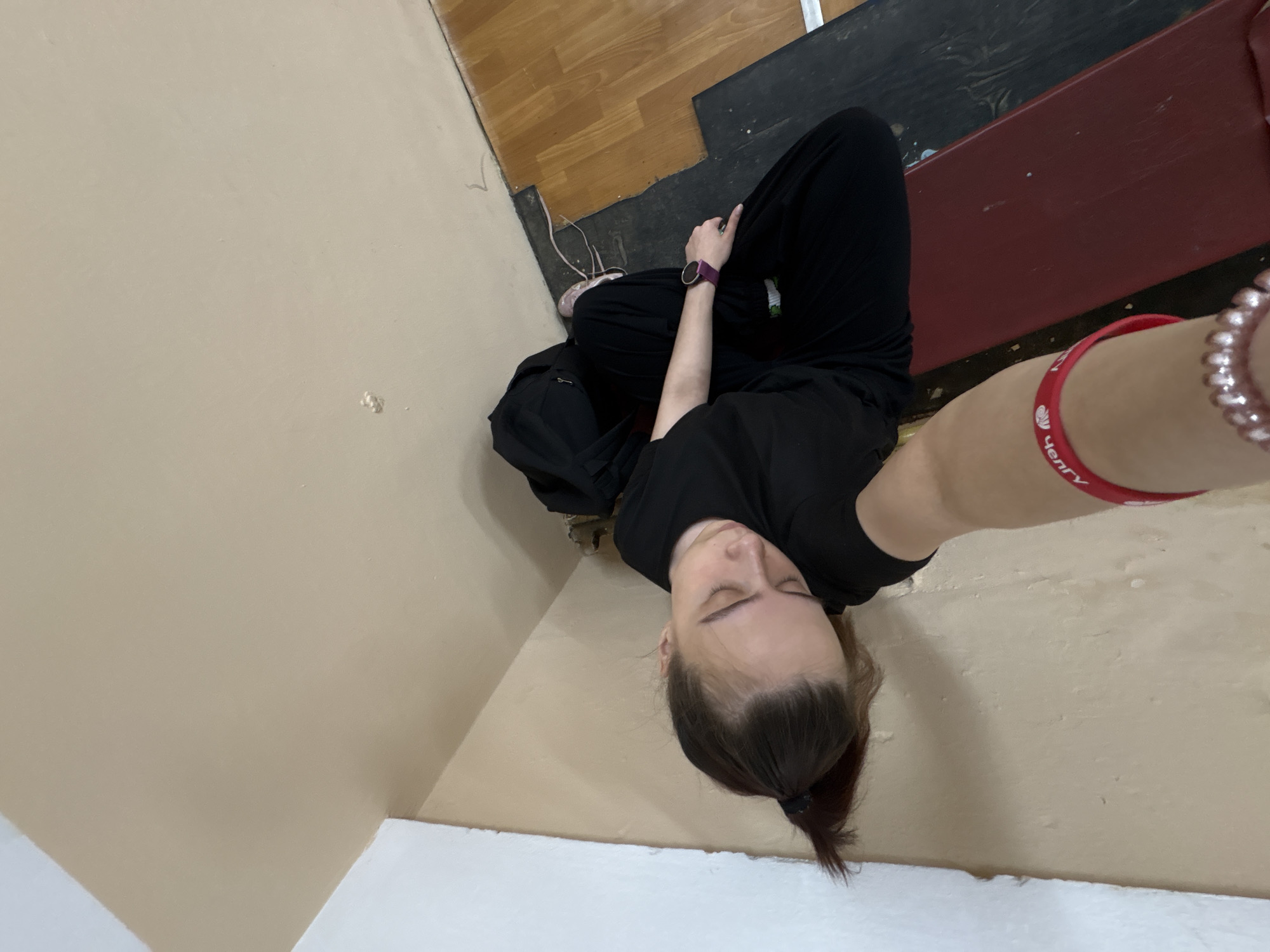 A selfie shows a person with brown hair tied back in a ponytail, wearing a black T-shirt and black pants, sitting cross-legged. The person is also wearing a wristwatch and bracelets. The background includes a wall painted beige, a wooden floor, and a red bench. The red bracelet has "челгу" written on it, which suggests that the person is located in or near Chaikovsky, Russia.