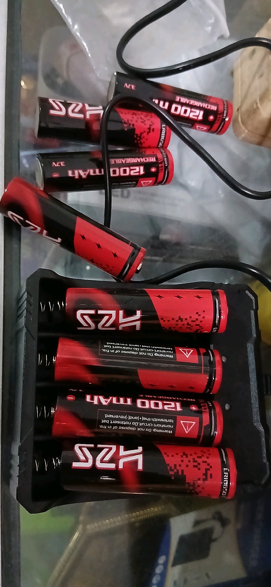 An overhead, eye-level medium shot captures a collection of rechargeable batteries and a charger. The batteries are red and black with white lettering indicating "HZS" and "1200 mAh RECHARGEABLE." One battery is positioned horizontally in the foreground, angled towards the left, with its positive terminal visible. Behind it, a black battery charger holds four more batteries vertically, all facing upwards. The charger itself is black and rectangular with textured sides. Two additional batteries, similar to those in the charger, are seen at the top of the frame, one horizontally placed and partially obscured by a black cable, and another in the upper right, also partially hidden. The background is blurred, suggesting an indoor setting with reflective surfaces. There are no people visible in the image. The scene does not provide any information to associate it with Samarinda, Indonesia.