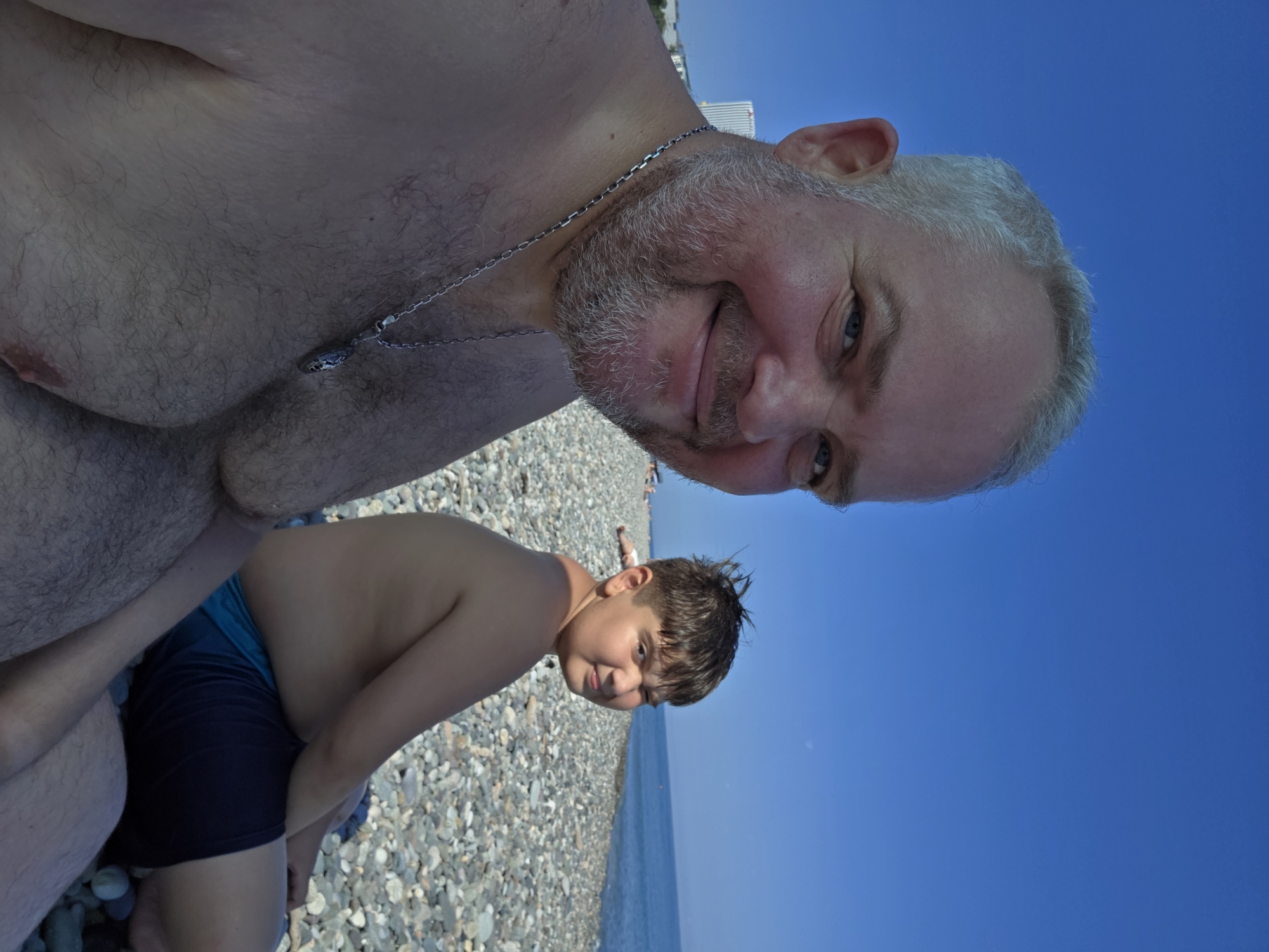 An outdoor, medium shot captures two males on a pebble beach under a clear blue sky in Batumi, Georgia. The man in the foreground, closer to the camera, is older with graying hair and a beard, wearing a silver chain. He is looking towards the camera with a slight smile. To his right and further back, a younger male with dark hair is also looking towards the camera, with one eye squinted, wearing blue swim trunks. The background shows a body of water, likely the Black Sea, and a building in the distance. The ground is covered in small, grey pebbles.