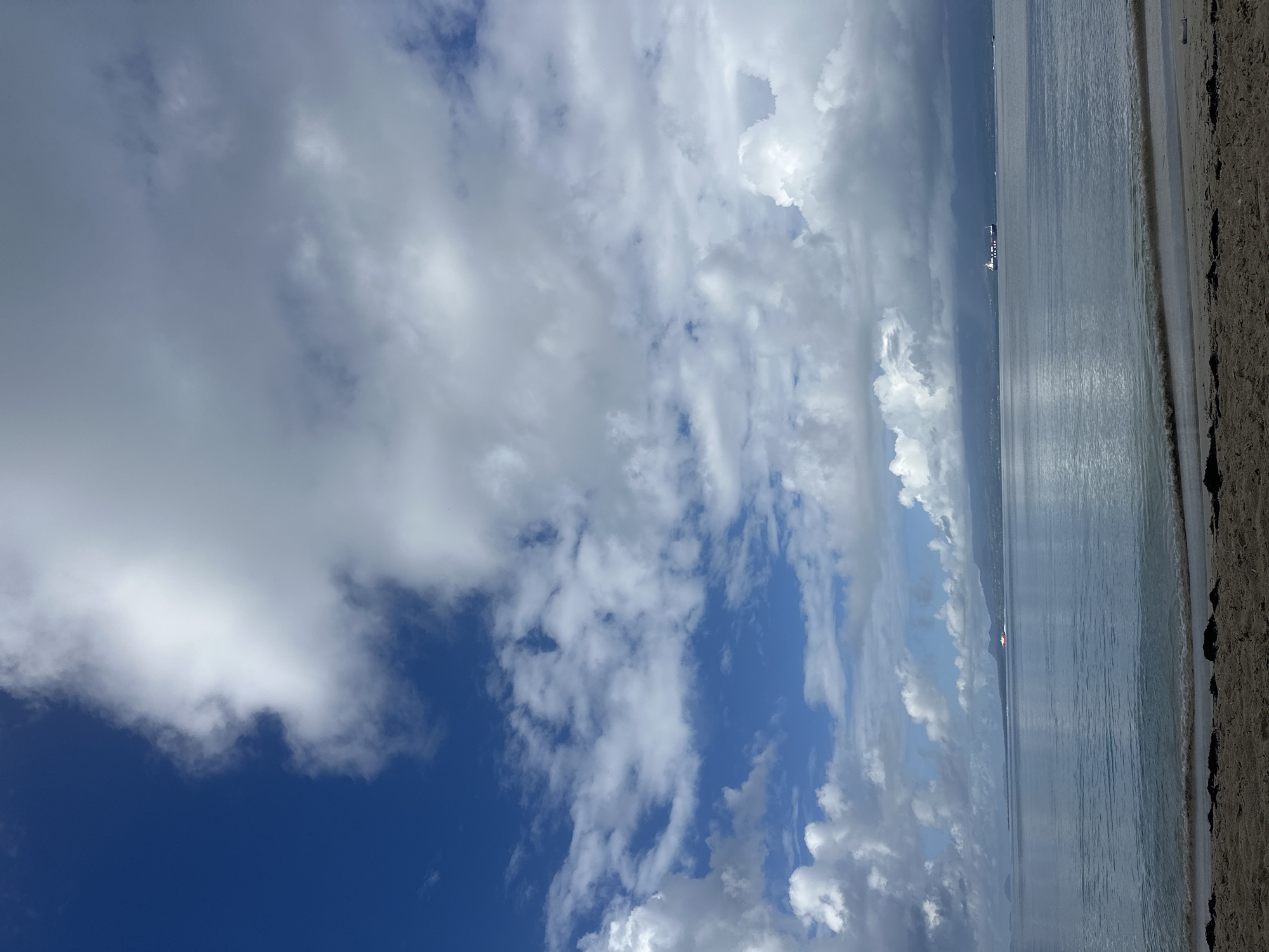 The image shows a view from a beach in Le Gosier, France. The sky is partially covered with cumulus clouds, with patches of blue visible. The ocean stretches out to the horizon, with a line of land visible in the distance. A large cargo ship appears on the water further from the beach. The beach itself is sandy and stretches along the right edge of the frame. A gentle wave is visible breaking on the shore. The image is taken during the daytime with natural lighting. The perspective is from a low angle, looking out towards the ocean and sky.