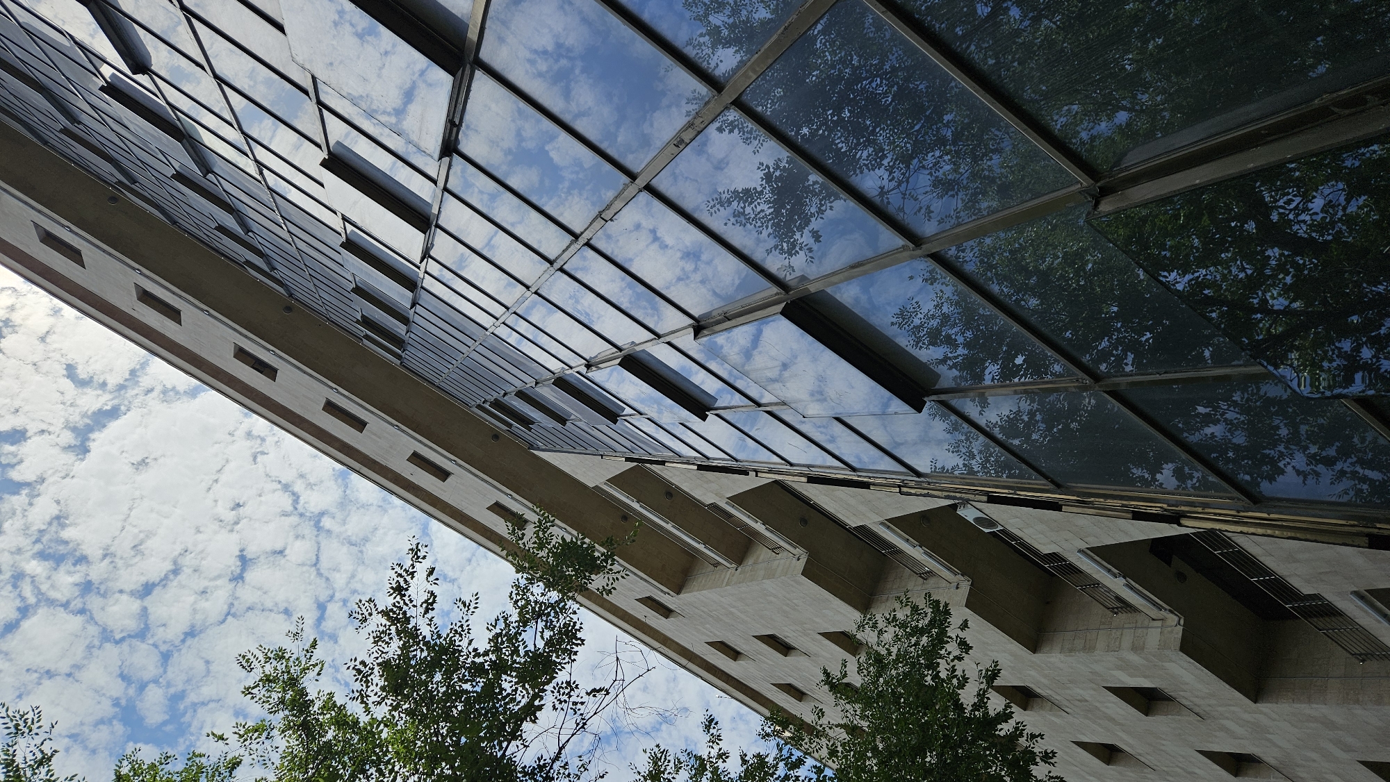 The image captures a low-angle view of a modern building with a facade composed of beige stone and large glass panels. The building appears to be located in Almaty, Kazakhstan, as indicated by the architectural style and the presence of trees that are common in the region. The glass panels reflect the blue sky, white clouds, and the surrounding green trees. The stone facade features a series of rectangular windows and balconies with metal railings. The sky is partly cloudy, with scattered white clouds against a bright blue background. The overall scene is one of urban architecture integrated with natural elements.