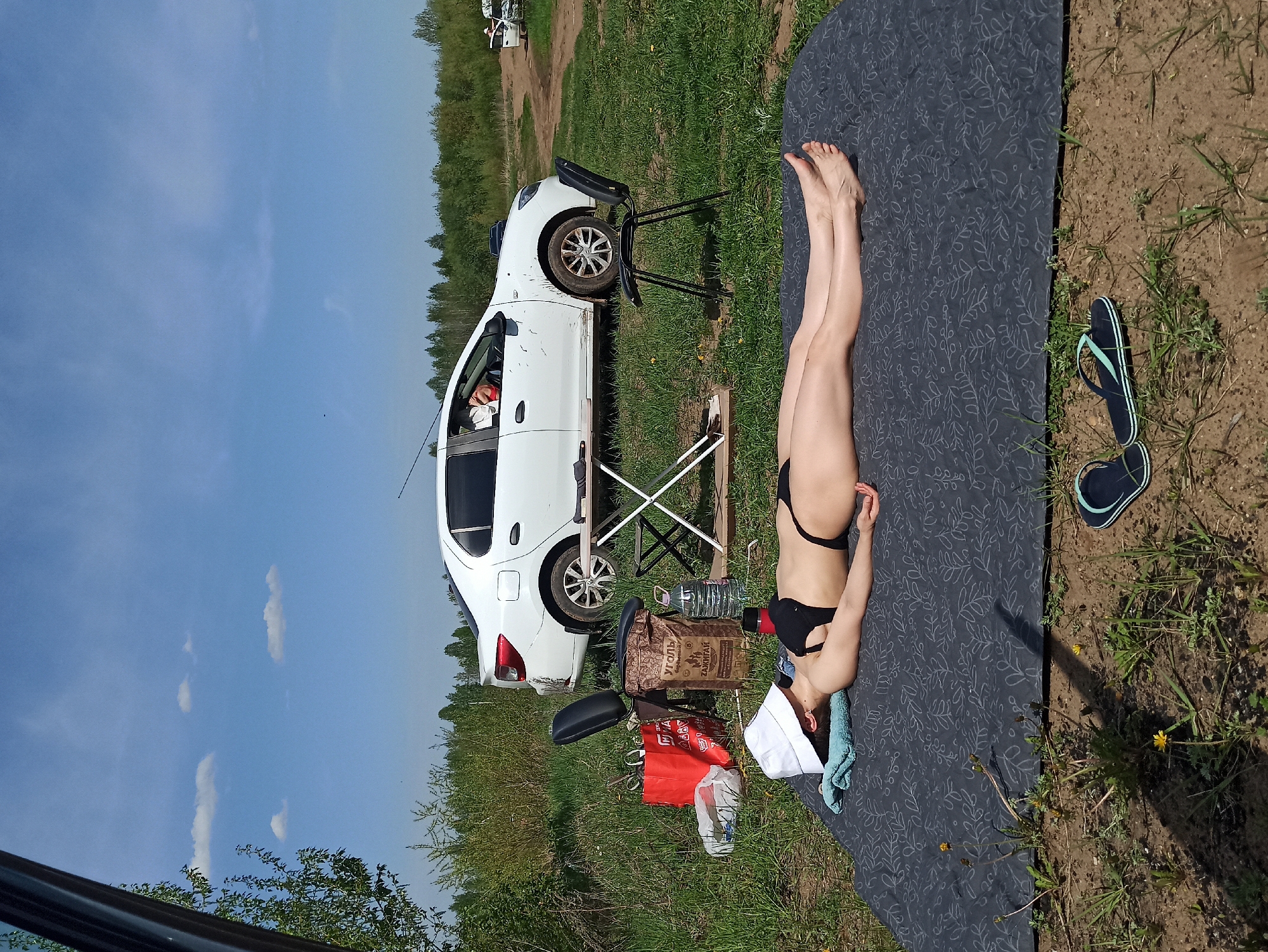 A woman wearing a black bikini lies on a patterned grey mat in a grassy area next to a white car. The car is slightly elevated by a black chair under one of its wheels. A folding table and a bag of charcoal are near the car. Flip-flops are on the ground next to the mat. The scene is set outdoors, with trees visible in the background and a partly cloudy sky overhead.