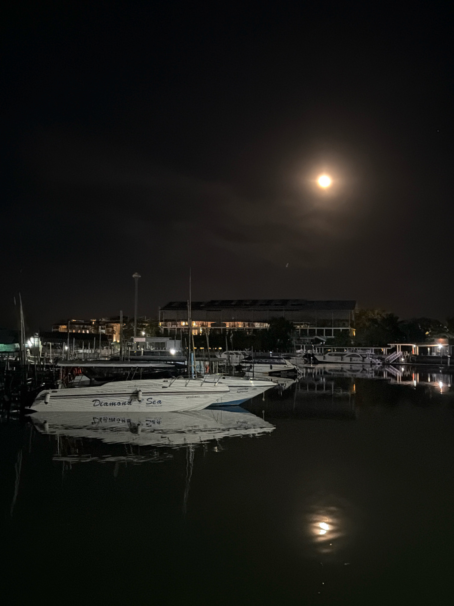 The image depicts a nighttime scene at a harbor in Ko Kaeo, Thailand. The sky is dark with a full moon visible in the upper right quadrant, its light illuminating some of the clouds. A prominent white boat, named "Diamond Sea," is docked in the foreground, its name clearly visible in blue lettering. The boat is reflected in the dark, still water. Several other boats are docked behind it, and a building with illuminated windows can be seen in the background, along with palm trees. The overall atmosphere is calm and serene, illuminated by the moonlight and the artificial lights from the buildings and boats. There are no people visible in the image.