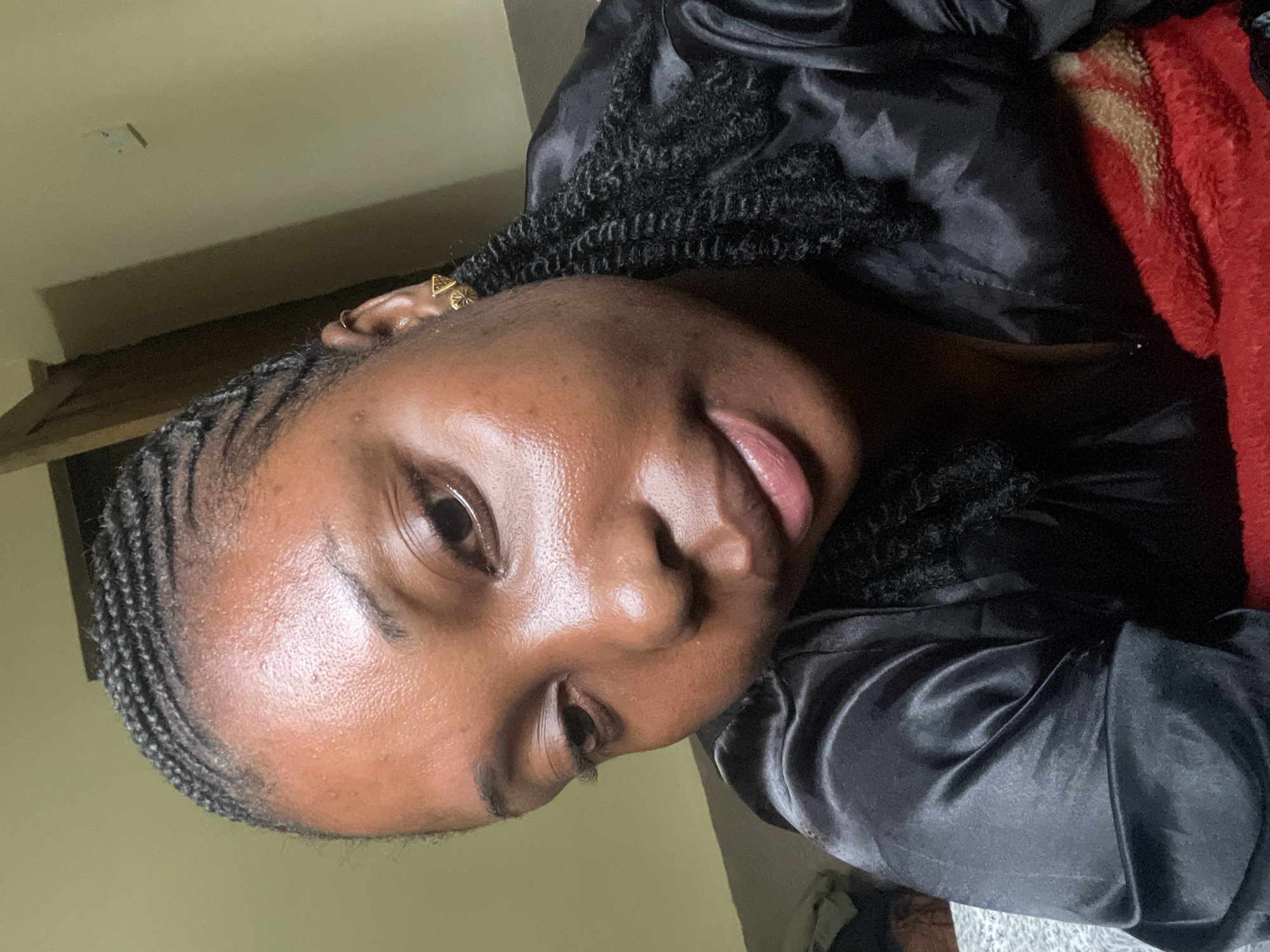A close-up portrait of a Black woman with braided hair is presented. The woman is wearing earrings in her left ear. She is attired in a black satin garment. Part of a red blanket is visible in the upper right. The background is a yellow-toned wall. There are no indicators to discern the location as Jos, Nigeria.