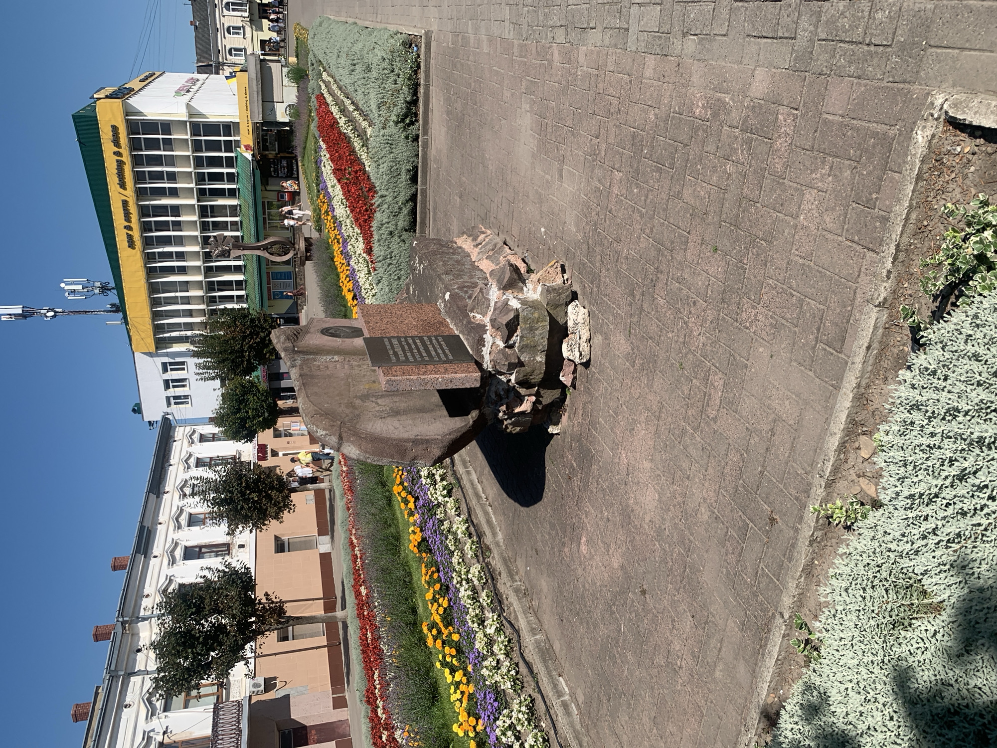 The image depicts a paved public square in Kolomyia, Ukraine, on a clear, sunny day. In the foreground, a large, weathered stone monument is centrally located, appearing to be a historical marker or memorial. It is composed of rough-hewn rocks at its base, topped with a more polished stone slab bearing inscriptions and a relief image.
Behind the monument, a meticulously arranged flower bed displays a vibrant array of colorful flowers, arranged in diagonal stripes of red, white, yellow, and purple, interspersed with lush green foliage. This flower bed runs along a pathway.
Further in the background, a multi-story building with a yellow and green facade dominates the scene. The building features numerous windows and has signage that reads "new & espem / clothing & shoes." Pedestrians are visible walking in front of this building and near the flower beds. To the left of the building, a white building with red-tiled roofs and decorative architectural elements can be seen.
The ground is paved with interlocking brick pavers, characteristic of public pedestrian areas. The sky is a clear, vibrant blue, indicating fair weather conditions. The overall impression is of a well-maintained urban public space with a historical monument and floral displays.
