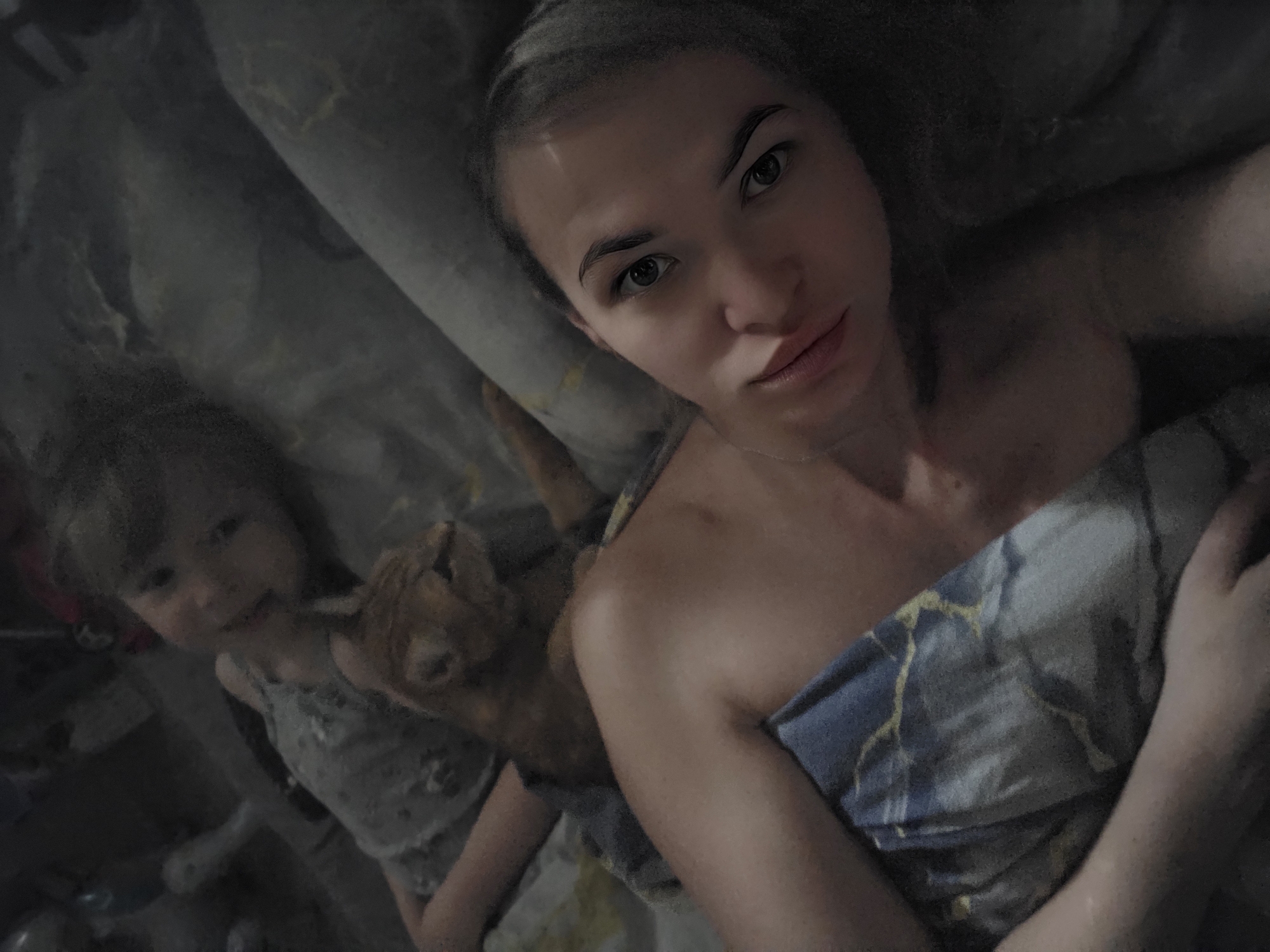An intimate, dimly lit indoor scene captures a woman, a young child, and a small goat kid lying together, likely in a bed, in Chaikovsky, Russia. The image is taken from a high angle, appearing to be a selfie by the woman who is in the foreground. She looks directly at the camera with a neutral expression, revealing light hair pulled back, and her bare shoulders above a blue blanket adorned with a distinctive golden, abstract, or cracked pattern.
To her left, slightly further back and out of focus, a young child with darker hair is visible, wearing a light-colored top. Her expression is calm. Nestled between the woman and the child is a small, light brown goat kid, also lying down. The animal's head and part of its body are visible, adding an unusual and tender element to the scene.
The overall lighting is low, suggesting either early morning, late evening, or an interior with subdued illumination. The bedding appears to be in shades of grey. There is no visible text in the image. The atmosphere conveys a sense of quiet togetherness and relaxation.