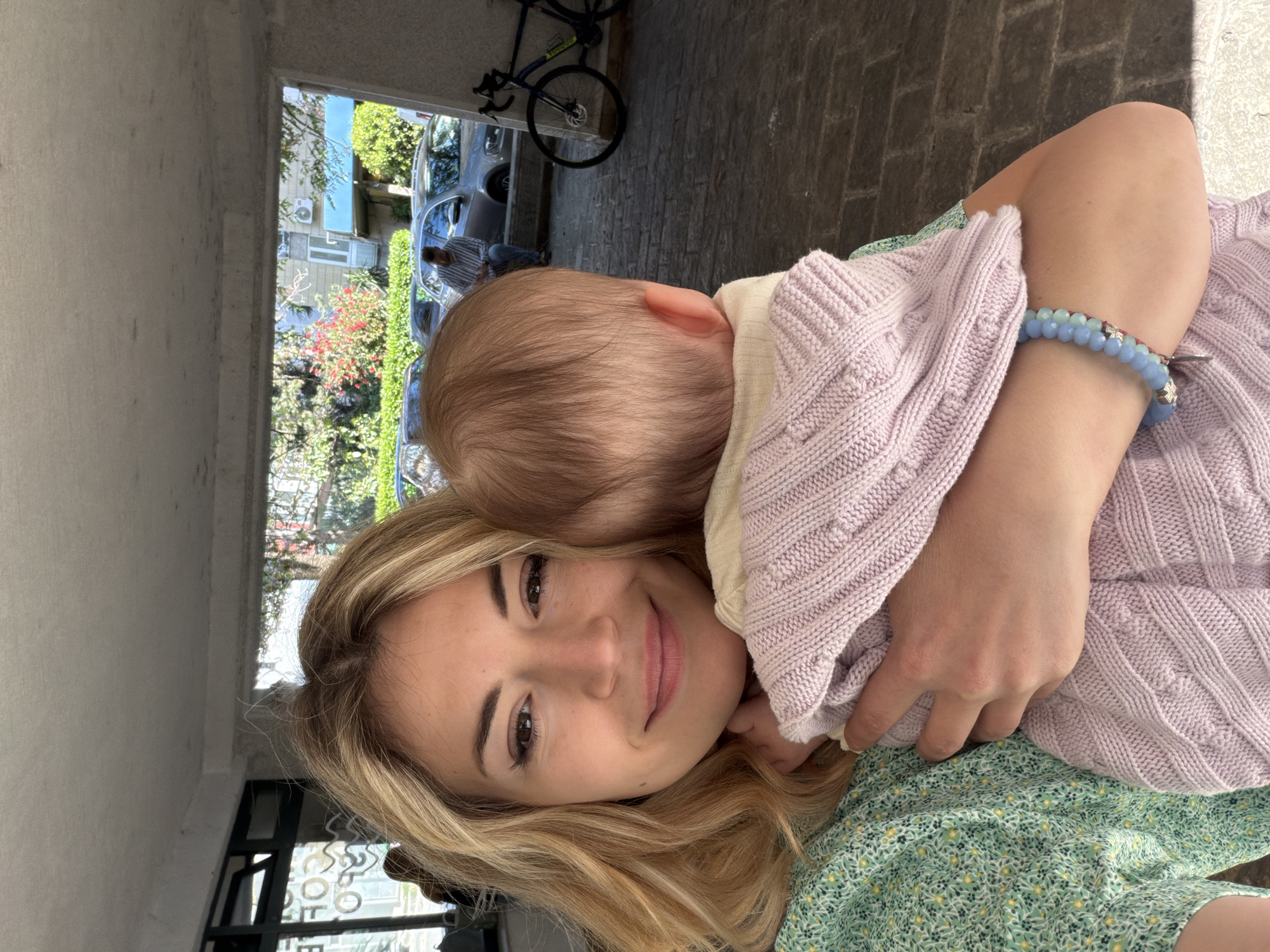 The image captures a selfie-style portrait of a woman holding a baby, taken outdoors during the daytime. The woman, with long blonde hair, a bright smile, and dark eyes, looks directly at the camera. She wears a light green dress with a small floral pattern, and a blue beaded bracelet is visible on her right wrist. She is embracing a baby whose back is to the camera, with only the light brown hair on the back of its head and right ear visible. The baby is wrapped in a light purple knitted blanket or sweater and appears to be resting against the woman's shoulder.
They are positioned under a covered structure or archway, with bright sunlight illuminating the outdoor scene behind them. The ground is paved with stones or bricks. Through the opening, a vibrant background reveals lush green trees and bushes, some with red flowers, suggesting a warm climate. Parked cars are visible, and a person in a striped shirt stands nearby. In the upper right background, a dark bicycle, possibly a "SPECIALIZED" brand, leans against a wall. Parts of buildings, including one with an air conditioning unit, are also visible. The woman's expression conveys happiness and affection. Faint, blurry text, possibly "OFFICE," can be seen on a glass surface in the lower left background. The setting, with its stone pavement and greenery, is consistent with an urban or suburban area like Budva, Montenegro.