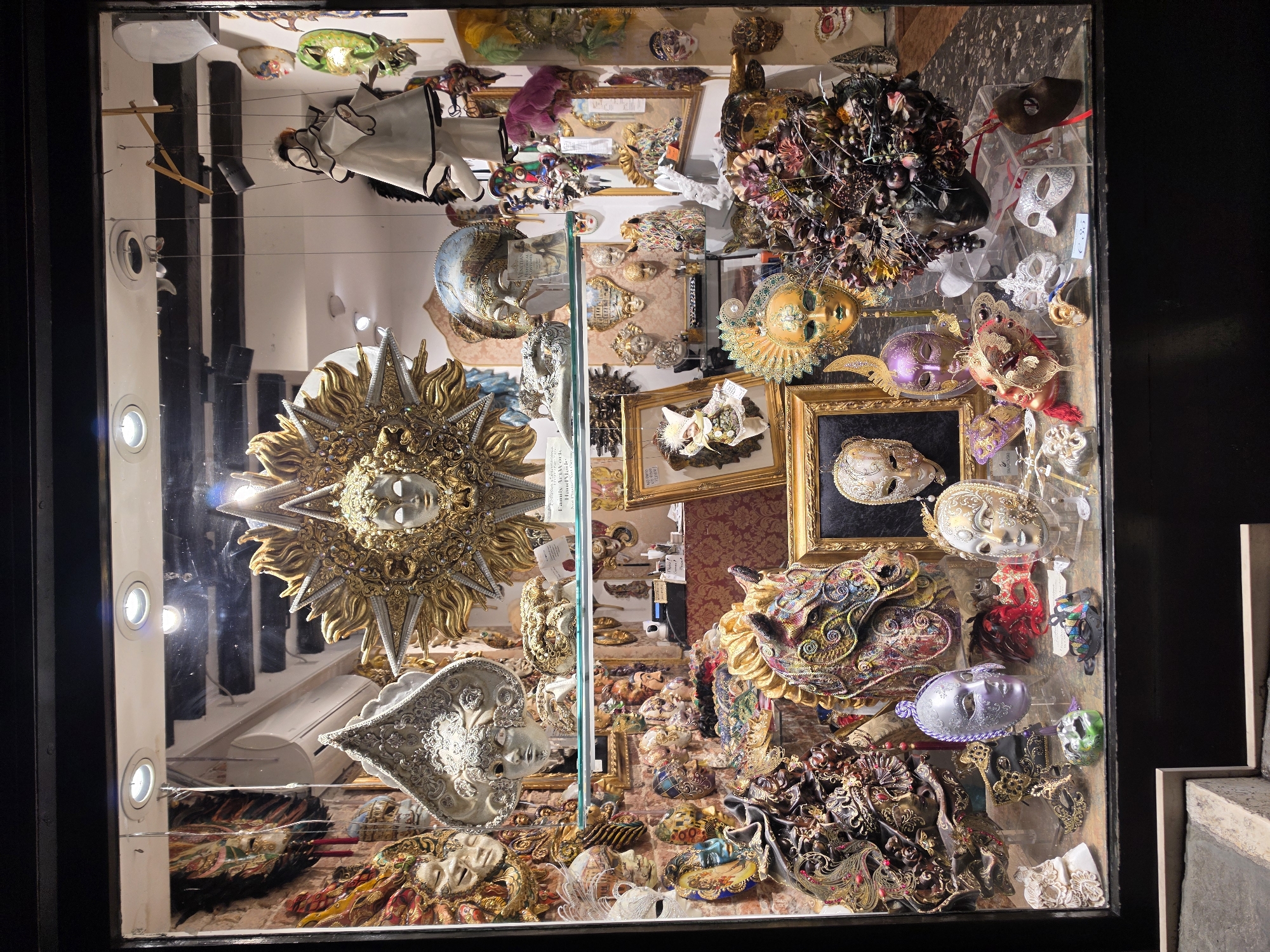 This is a photograph of a Venice mask shop window display, likely located in the San Marco district of Venice, Italy, given the abundance of ornate and traditional Venetian masks. The display is densely packed with a variety of masks, primarily of the Venetian style, made from materials such as porcelain, plaster, paper-mâché, and adorned with feathers, glitter, and intricate paintings.
In the upper section of the window, a large, elaborate sun-shaped mask with a central human face is prominently featured. To its left, a white ornate mask with silver detailing is visible, and to its right, a marionette dressed in a white and black outfit hangs by strings. Various other masks, including those with animal motifs like a horse's head and more abstract designs, are displayed on shelves and hung on the walls.
The lower section of the display showcases a further assortment of masks in various sizes and colors, including a shimmering purple mask, a gold and green mask with feathered headdress, and a striking black mask adorned with a dense arrangement of flowers and fruits. Smaller, simpler masks and decorative items are also present, some with price tags. The overall impression is one of opulence and a celebration of Venetian carnival traditions. The lighting within the display creates a bright, spotlighted effect on the masks. The shop window is framed by a dark, possibly wooden, structure.