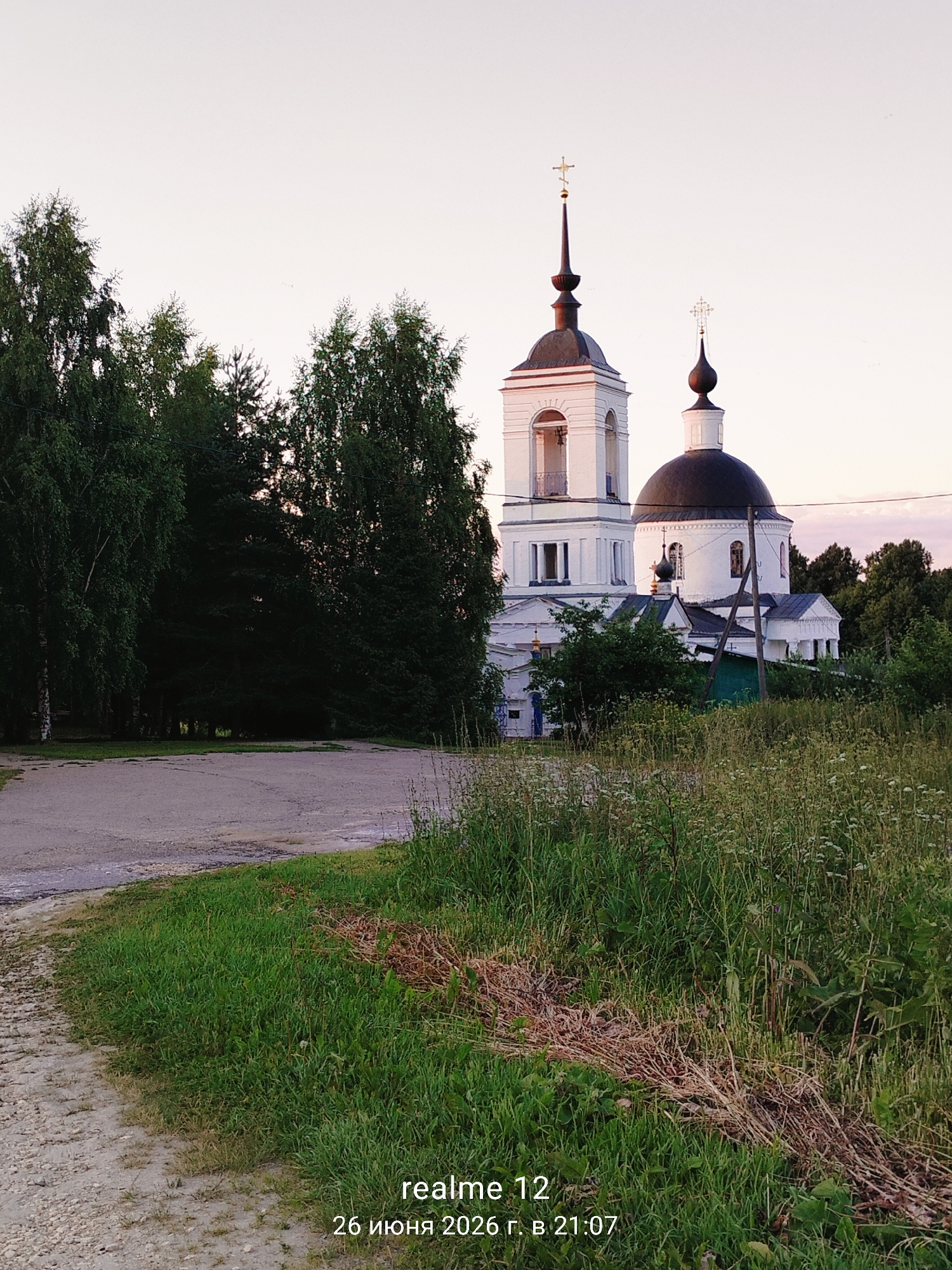 A white Orthodox church with a dark dome and a bell tower stands amidst lush green trees. The church has multiple arched windows and ornate crosses atop its spires. A dirt path leads towards the church from the lower left of the frame, bordered by tall grass and wildflowers. The sky is a pale pink and white, suggesting either dawn or dusk. The scene is peaceful and rural, likely in Novoe, Russia, given the timestamp "26 июня 2026 г. в 21:07" which translates to "June 26, 2026 at 21:07". There are no people visible in the image.