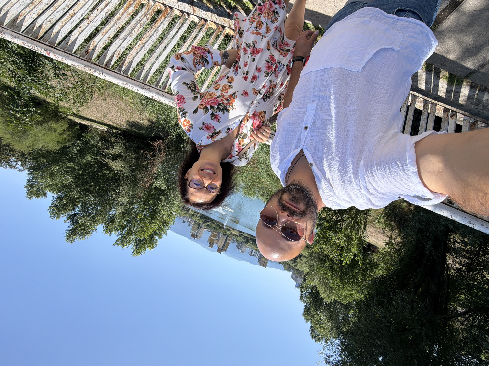 A man and a woman are taking a selfie outdoors on a bright, sunny day. They are standing on a metal railing that appears to be part of a bridge or walkway. The man is on the left, wearing a white linen shirt and sunglasses, with a bald head and a beard. The woman is on the right, wearing a white floral dress and glasses. Behind them, there is a body of water, likely a lake, with a fountain in the middle. In the background, there are several tall, modern buildings with a golden facade, typical of the city of Sofia, Bulgaria. The surrounding landscape consists of lush green trees and vegetation, with mountains visible in the distance. The railing shows signs of rust, indicating it may be an older structure. The overall scene suggests a pleasant day spent by the lake in a city park.