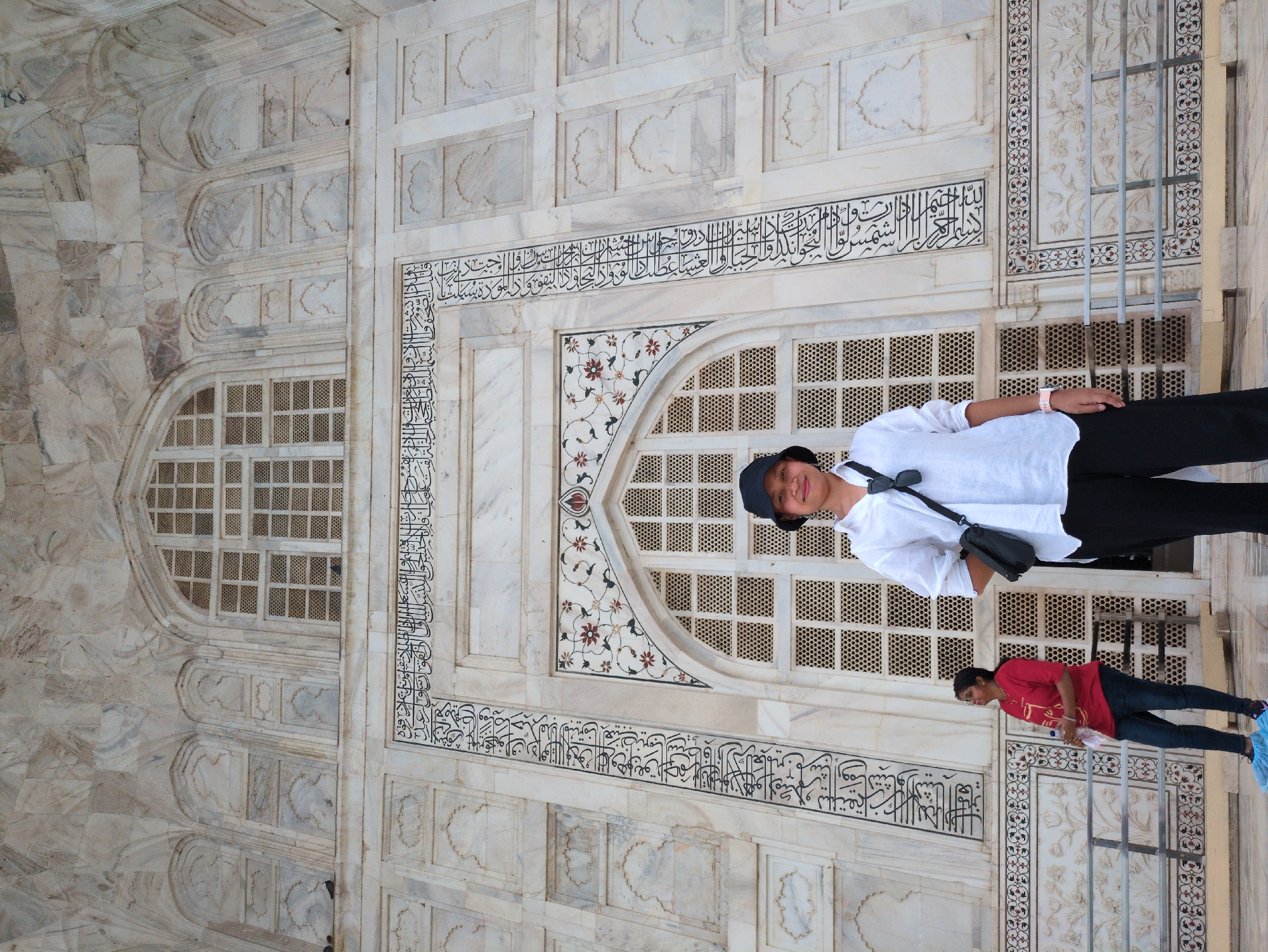 A full-body, low-angle outdoor shot shows a woman standing in front of a large, ornate marble building with intricate carvings and latticed windows, likely the Taj Mahal in Agra, India. The woman, positioned in the center foreground, wears a white collared shirt, black pants, and a black bucket hat. She has a black crossbody bag slung over her shoulder. To her left, a woman in a red t-shirt and blue jeans stands looking upwards and holds a clear plastic bottle with a red label. She is wearing blue shoe covers. The building's facade is predominantly white marble with decorative black calligraphy and floral motifs. Metal railings are visible in the foreground. The sky is overcast, providing soft, diffused lighting.