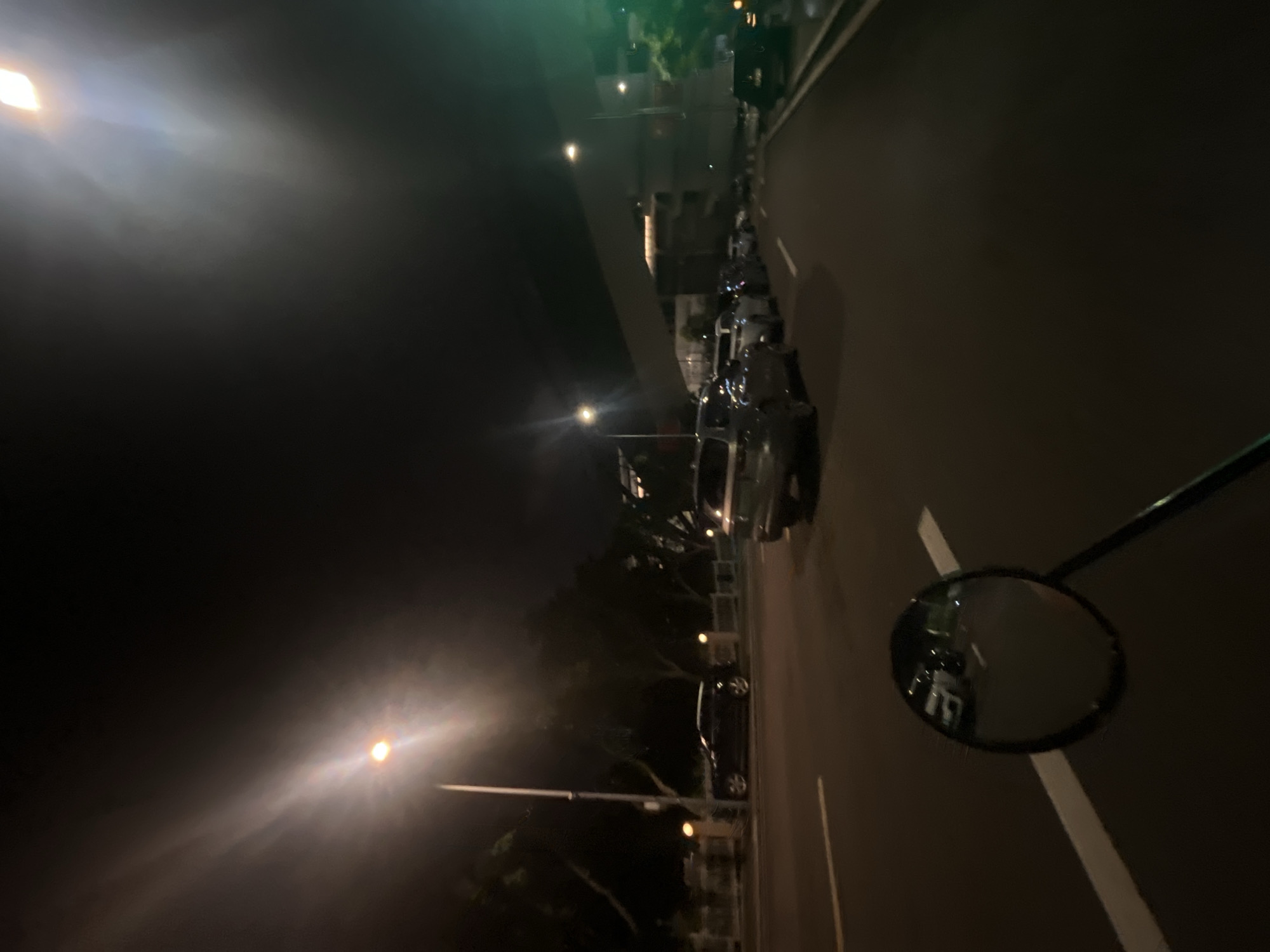 The image depicts a nighttime scene on a road in the Jiulongpo District, China. The perspective is from a vehicle, likely a motorcycle, as evidenced by the circular rearview mirror in the foreground reflecting a portion of the road and traffic. The road is paved and marked with white lines. On the right side of the road, several cars are parked in a line. On the left side of the road, there is a sidewalk or median with trees and lampposts. In the background, a modern building with illuminated windows is visible, and a prominent light fixture with a bright glare illuminates the upper portion of the image. The overall atmosphere is dark, with streetlights and building lights providing the primary illumination. There are no people clearly visible in the image.