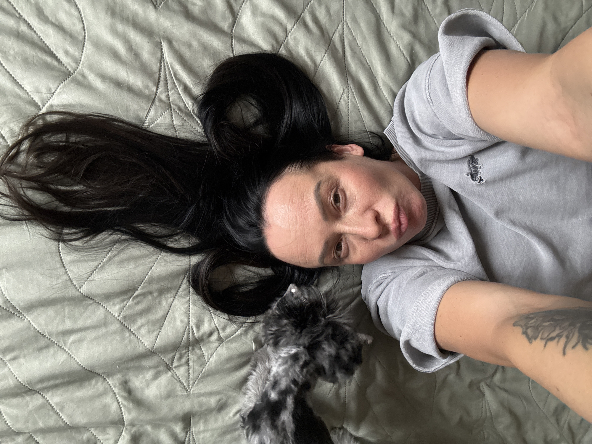 A woman with long dark hair lies on a light green quilted surface, possibly a bed or sofa. She is wearing a light gray sweatshirt. Her hair is spread around her head. She is looking at the camera. A small, black and white dog is near her face. The location is not discernible from the image.