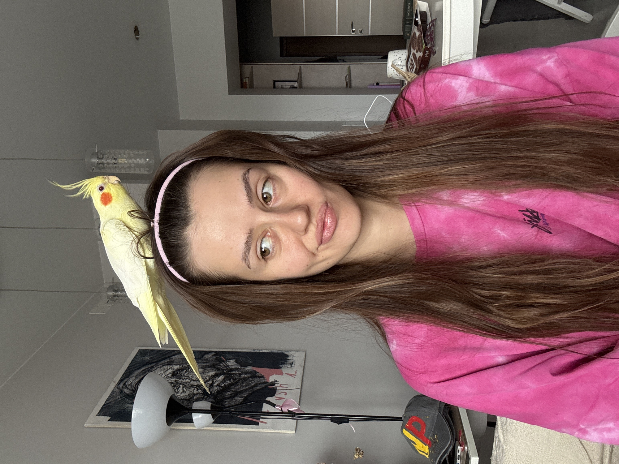 A woman with long brown hair and a pink headband is posing for a selfie indoors. A cockatiel is perched on her head. She is wearing a pink tie-dye sweatshirt. Behind her, there is a grey wall with a framed picture, a floor lamp, and a chandelier. Further back, a doorway leads to another room with shelves. The location is Krasnoyarsk, Russia.