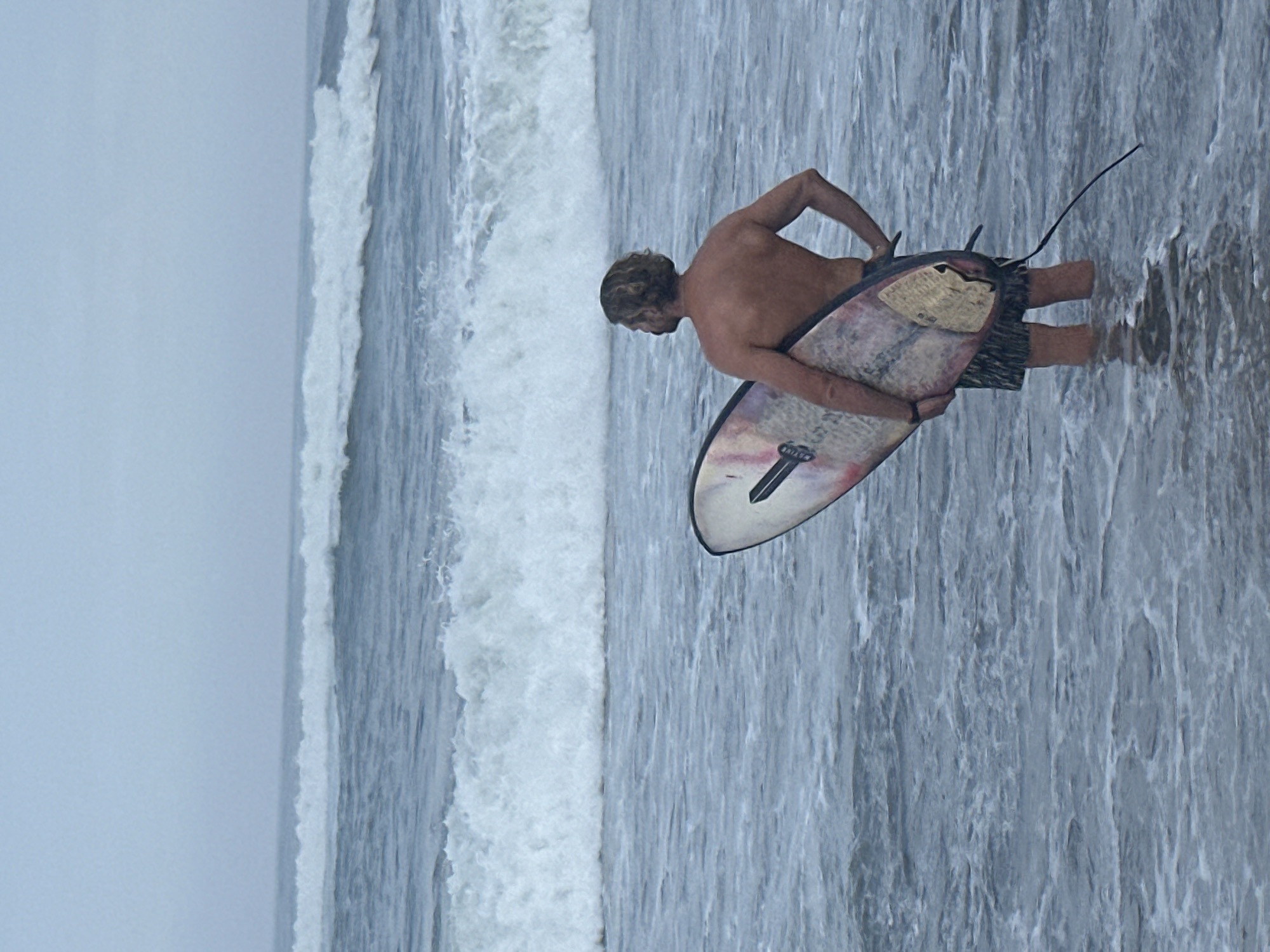 A man is standing in the ocean with a surfboard. He is facing away from the camera and looking out at the waves. The sky is overcast and the water is choppy. The man is wearing board shorts and is shirtless. The surfboard is multi-colored. This scene is likely at a beach in North Kuta, Indonesia, a popular surfing destination.
