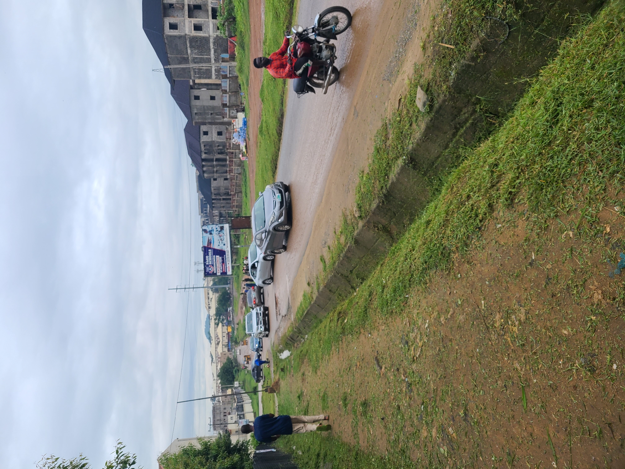 The image depicts a street scene in Abuja, Nigeria, under an overcast sky. In the foreground, a dirt path with sparse grass runs alongside a concrete drainage ditch. A single individual, a young man wearing a blue t-shirt and beige trousers, walks away from the camera on this path.
The street itself is wet, suggesting recent rain, and traffic is present. A man in a red shirt rides a red motorcycle, heading towards the viewer. Several cars are on the road, including a silver sedan and a grey hatchback. In the background, construction is evident with several multi-story buildings in various stages of completion, featuring concrete exteriors and dark roofs. A billboard advertising "Pace Schools" is visible. Power lines and poles are also present, along with distant hills. The overall environment appears to be a mix of residential or commercial development and undeveloped land.