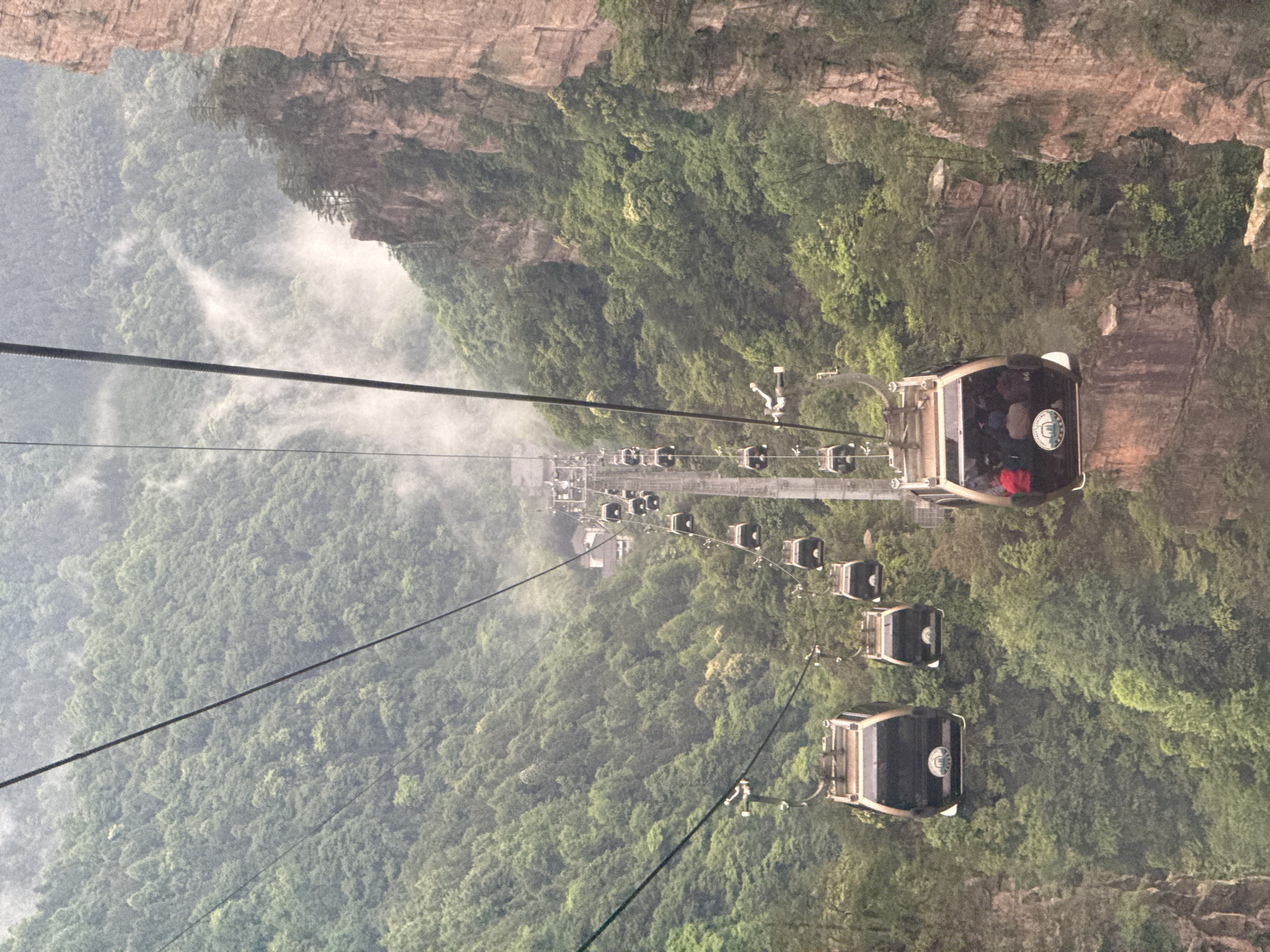 The image shows a cable car system in Wulingyuan, China. The cable cars are suspended over a lush, green valley. There are several cars visible on the cable, carrying passengers. The surrounding environment consists of trees, mist, and rocky cliffs.