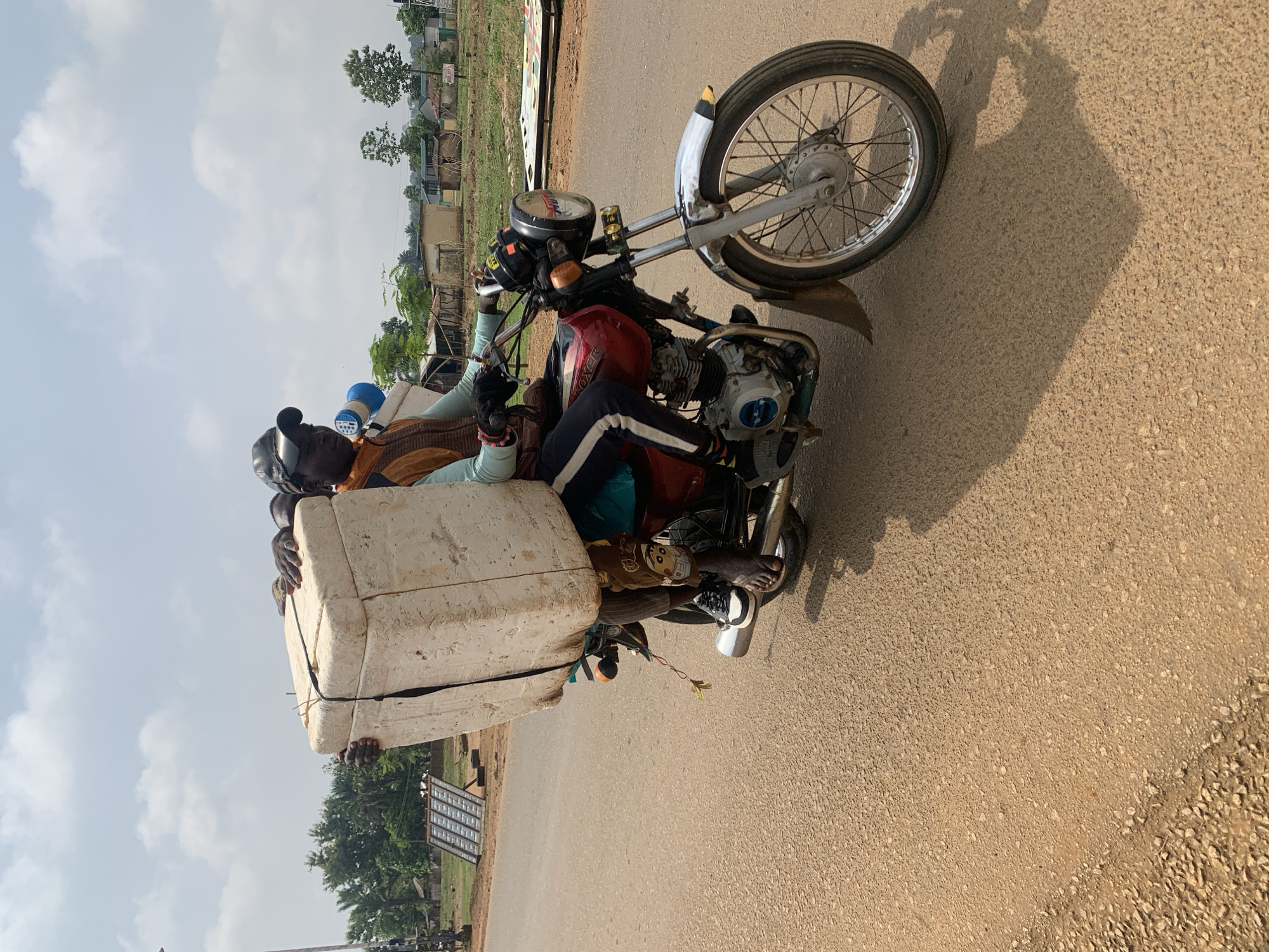 A heavily loaded motorcycle, viewed from a slightly elevated side angle, is captured on a paved, light brown road under a partly cloudy sky in what appears to be Maigizo, Nigeria. The image is oriented vertically (portrait), with the horizon line running along the right side.
The central subject is a red motorcycle with "BOXER" written on its fuel tank. It features chrome accents, spoked wheels, and visible engine parts, including a blue cap. A person, presumably the driver, is seated on the bike, wearing a brown and light-colored top, dark pants with white stripes, and black and white sneakers. A small blue container is perched behind the driver's head.
Attached to the rear of the motorcycle is a large, rectangular white object, possibly a cooler or a large block of foam, securely fastened with black straps. At least two other individuals or sets of hands are visible interacting with this large item; one pair of hands grasps the top, and another pair of bare feet and hands are visible near the bottom, suggesting people are either holding the load or also riding, partially obscured.
The bright sunlight casts distinct shadows of the motorcycle and its occupants onto the road, indicating it is likely mid-morning or early afternoon. The background features sparse greenery, trees, and several small, simple buildings, characteristic of a rural or semi-urban setting. The sky is bright blue with scattered white clouds. The scene portrays the practical transportation of goods and people on a motorcycle.