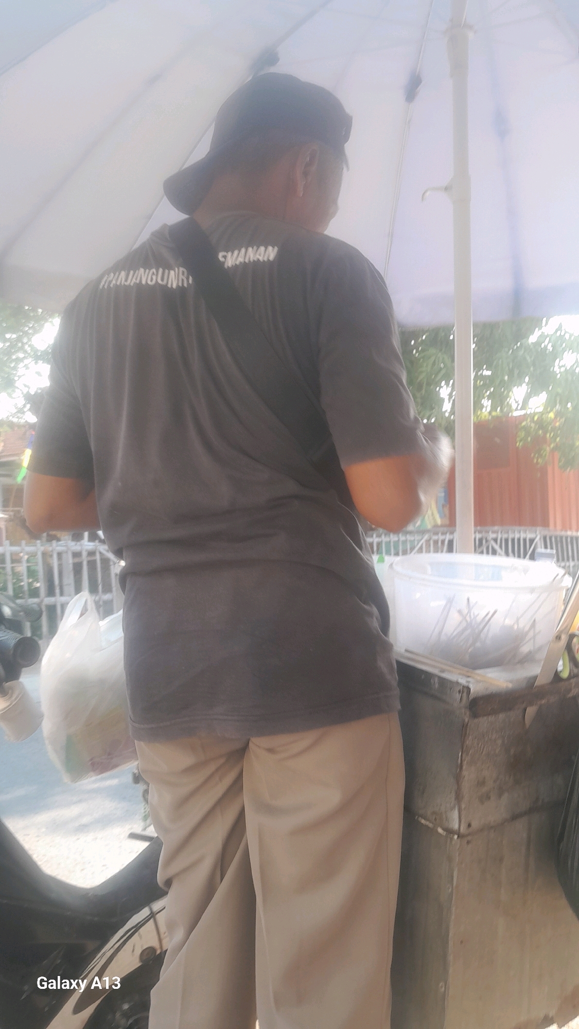 In Ngelo, Indonesia, a person stands from behind, facing away from the camera. They wear a dark gray t-shirt with white lettering that reads "#PANJANGUNR SMANAN" on the back and a black baseball cap. The individual is also wearing light brown pants. They are positioned next to a cart with a metal exterior, which holds a large clear plastic container filled with what appear to be eating utensils. A white plastic bag, possibly containing food items, rests on a motorcycle behind the person. The scene is partially covered by a large white umbrella, suggesting an outdoor setting, possibly a food stall or market. The background shows a glimpse of trees and a fence, indicating a moderately urban environment. The lighting suggests it is daytime, with a slight hazy quality, possibly due to the sun or atmospheric conditions.