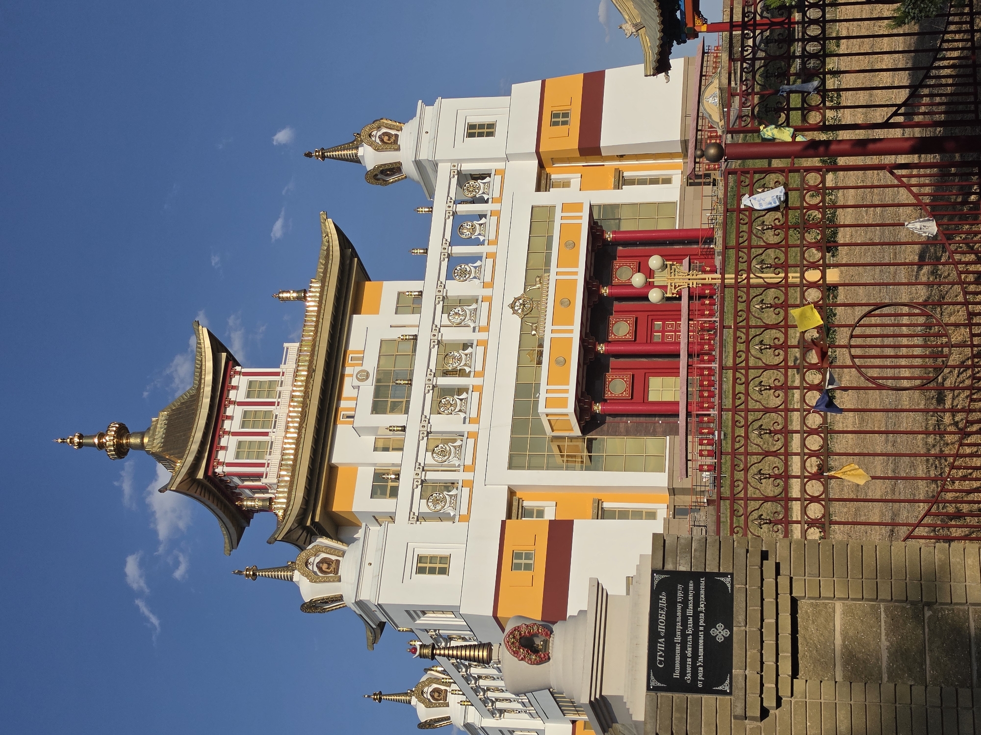 The image shows the "Golden Abode of Buddha Shakyamuni" Buddhist temple, also known as the central khurul of Elista, Russia. The temple is a large, multi-story white building with yellow and red accents, topped with gilded roofs and spires. The architecture is distinctively Buddhist, with prayer wheels and lotus motifs visible on the exterior. In the foreground, there is a brick fence with a metal gate, and a stone monument with a plaque that reads "STUPA 'VICTORY'" in Russian. Prayer flags are tied to the fence. The sky is clear blue with a few white clouds. The scene appears to be a bright, sunny day.