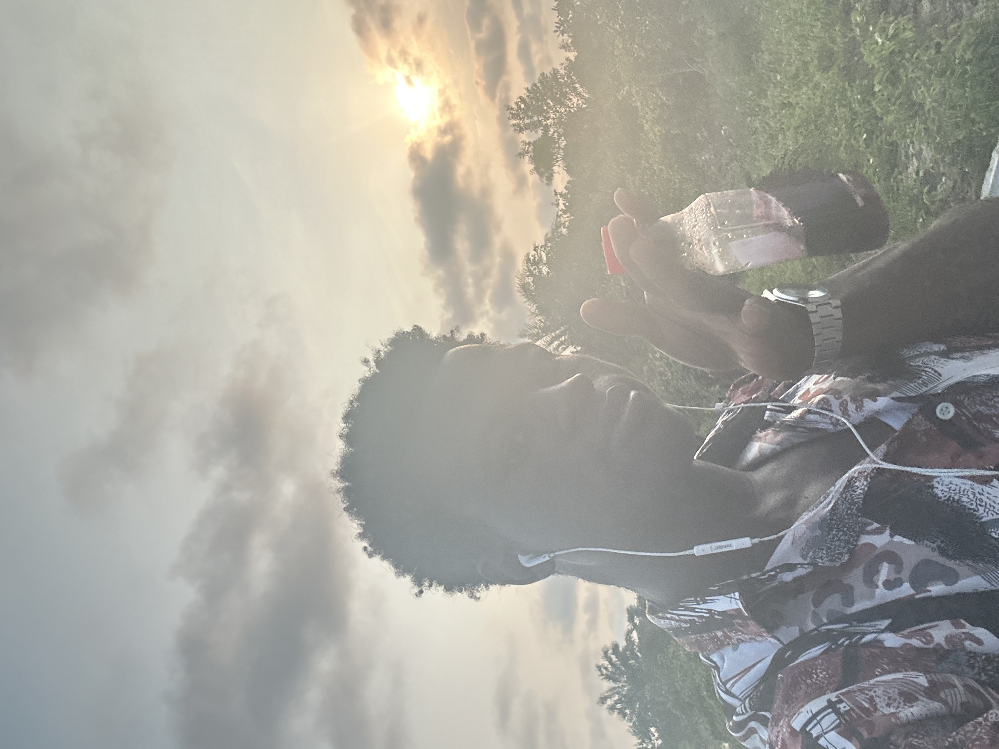 A selfie is taken by a young Black male in Badagry, Nigeria, with a hazy, golden sunset in the background. The man is wearing earbuds and a patterned shirt, and he holds a clear plastic bottle with a dark red liquid to his right. Trees and vegetation are visible in the distance, silhouetted against the bright sky. The overall atmosphere is warm and slightly diffused due to the sunlight.