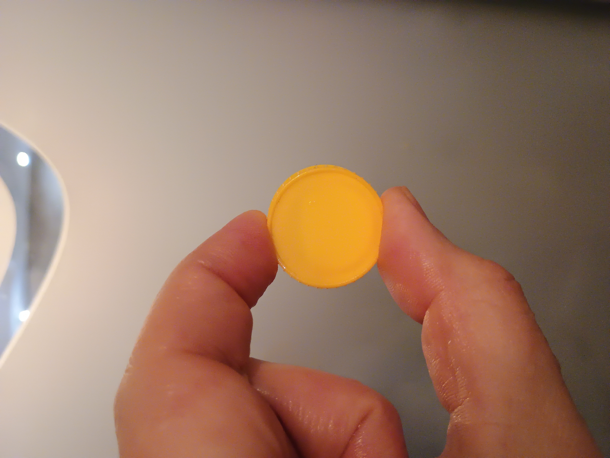 A close-up shot shows a person's hand holding a small, round, yellow object. The hand, with visible skin texture and some moisture, is positioned to the left of the frame, obscuring the background. The yellow object, which appears to be a cap or a button, is centrally located in the image and is the primary subject. The background is a plain, out-of-focus gray surface. The lighting is diffused, creating soft shadows. The location in Zaporizhzhia, Ukraine, is not discernible from the image.