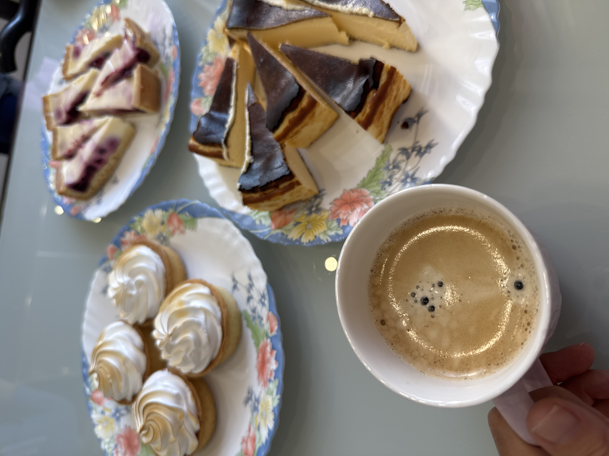 The image depicts a table setting with three plates of pastries and a cup of coffee. One plate holds slices of a burnt Basque cheesecake, the second contains small tarts topped with meringue, and the third features slices of a cake with a layer of jam. The coffee is in a white cup held by a person's hand. The pastries are arranged on floral-patterned plates. Given the style of pastries, the scene may be located in Saint Petersburg, Russia.