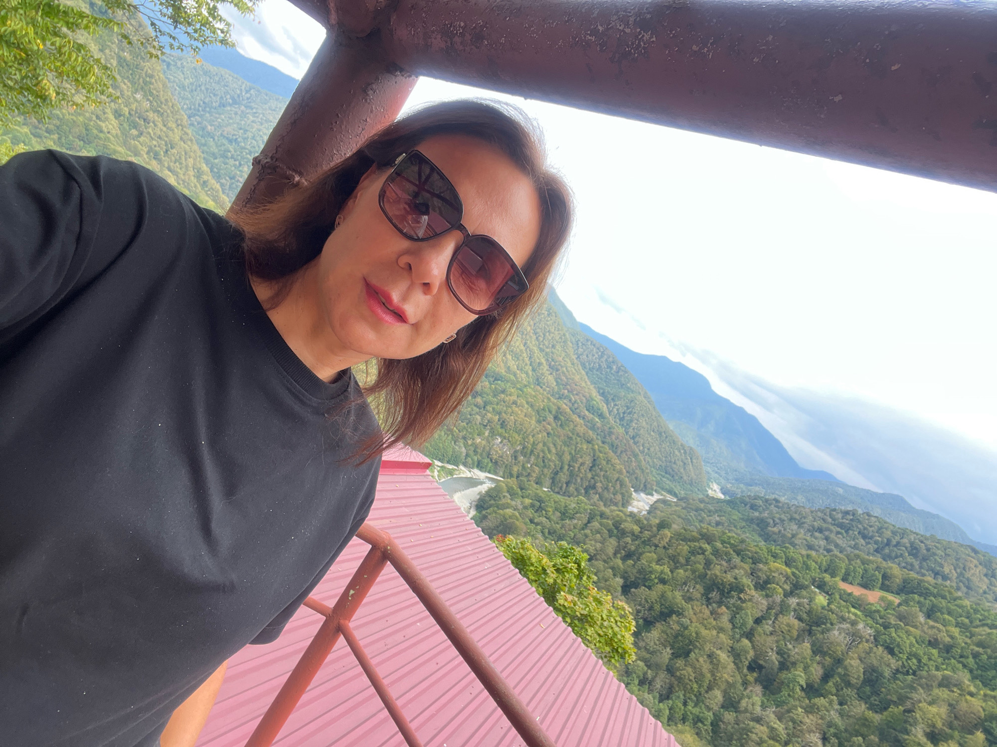 A woman with brown hair and sunglasses takes a selfie outdoors. She is wearing a black t-shirt and leaning against a red railing, with a mountainous landscape visible in the background. The mountains are covered in trees, and the sky is bright and overcast. A rusty metal beam is visible in the upper part of the frame. The woman is positioned in the foreground, with the scenic view behind her.